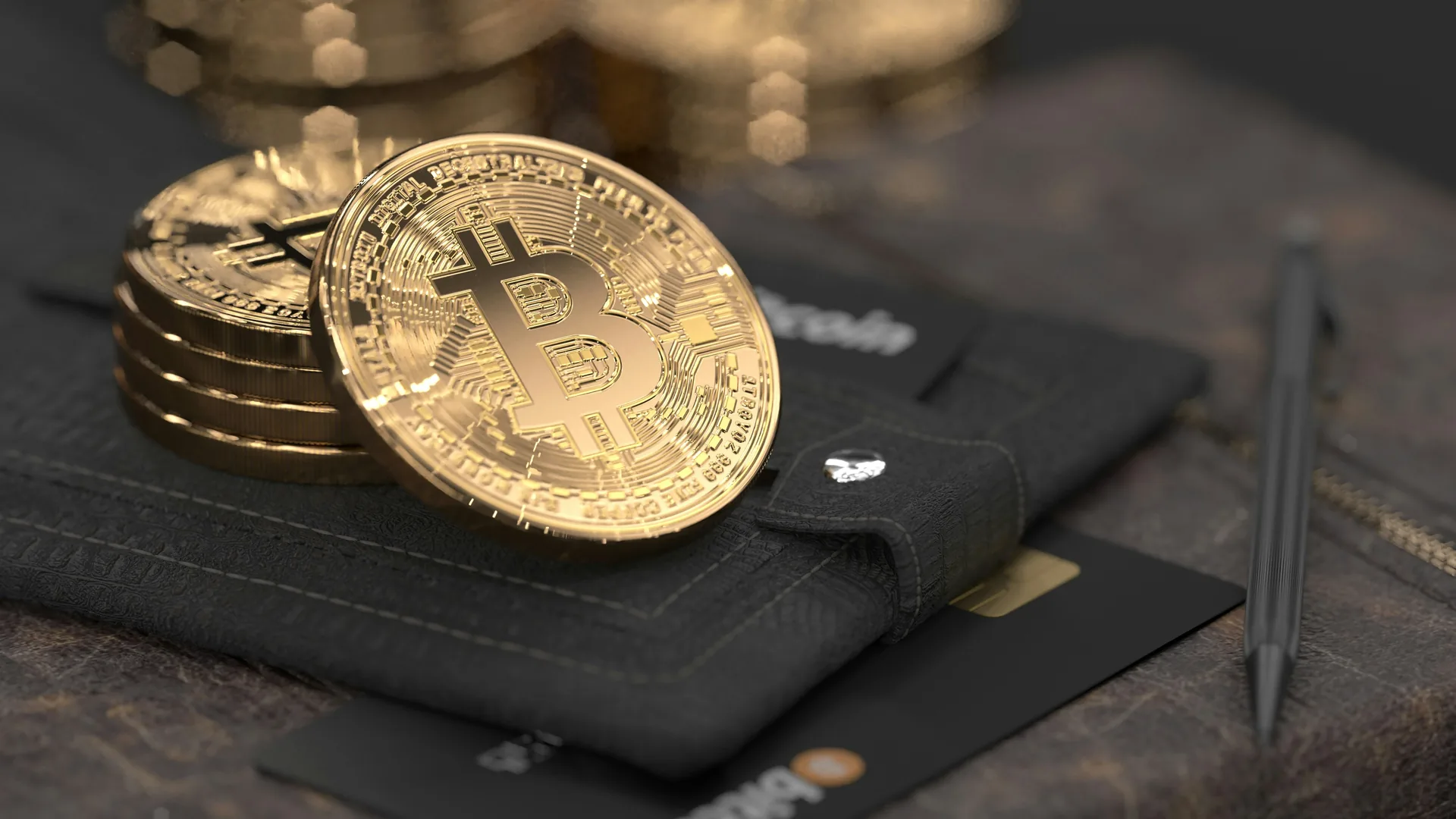 Gold Bitcoin coins resting on a dark leather wallet beside a pen