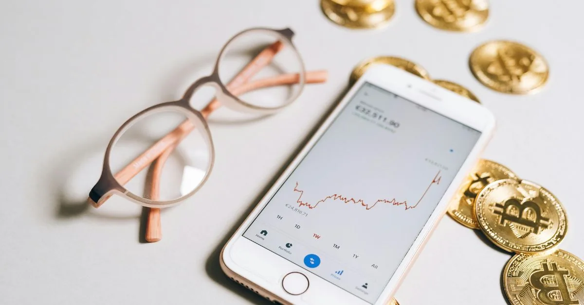 Phone showing a crypto price chart beside reading glasses and gold Bitcoin coins