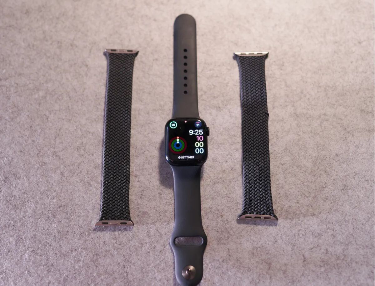 Apple Watch displayed alongside three interchangeable watch bands including a sport band and two braided solo loop options, laid flat on a gray surface