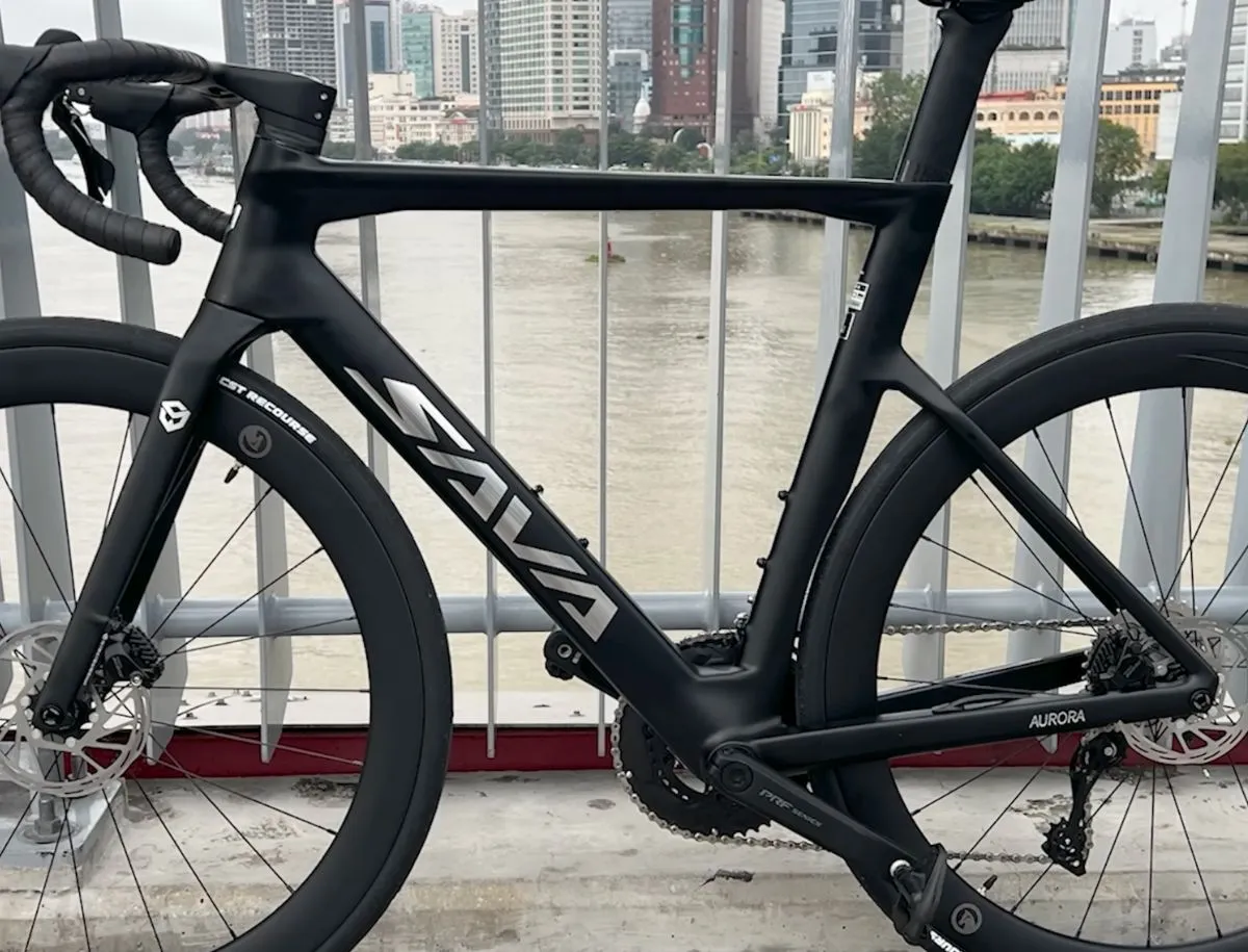 Close-up 3/4 profile of SAVA A7L Pro T800 carbon frame showing down tube SAVA logo, Aurora chainstay label, internal cable routing through the fork, CST Recourse tire, and Shimano hydraulic disc brake caliper, photographed on a bridge with city river backdrop