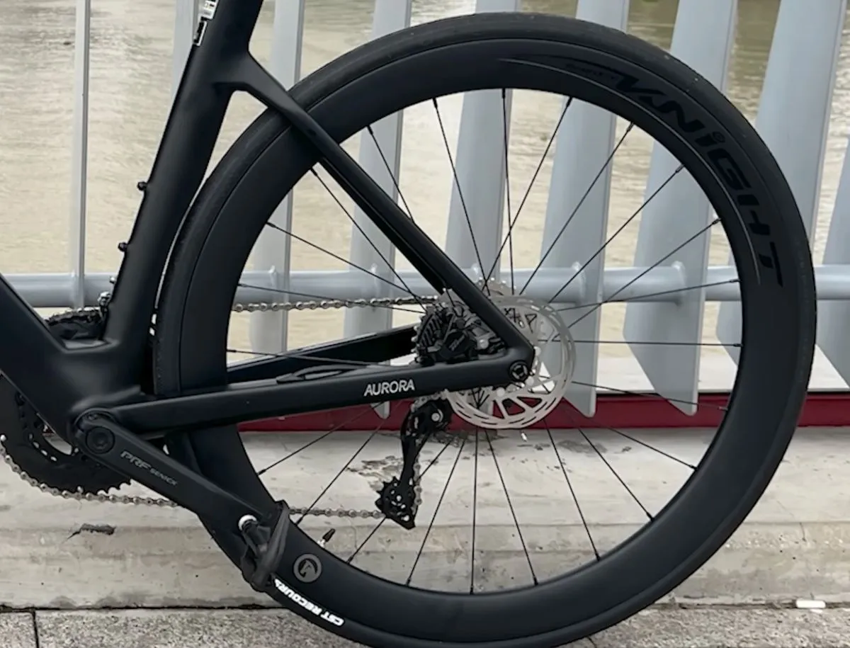 Rear view of SAVA A7L Pro showing deep-section carbon rear wheel with Knight branding, Shimano rear derailleur, disc brake rotor, and Aurora chainstay label, photographed beside a river