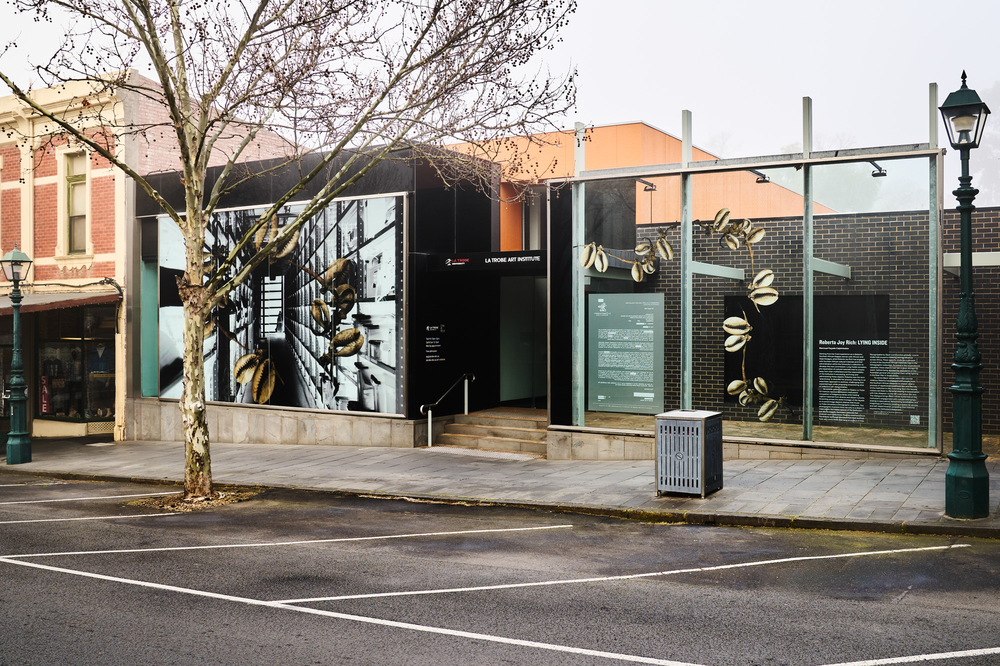 Street view of the gallery on a misty day, with the shell-and-shelving artwork in the main window and text panels on a glass structure to the right.
