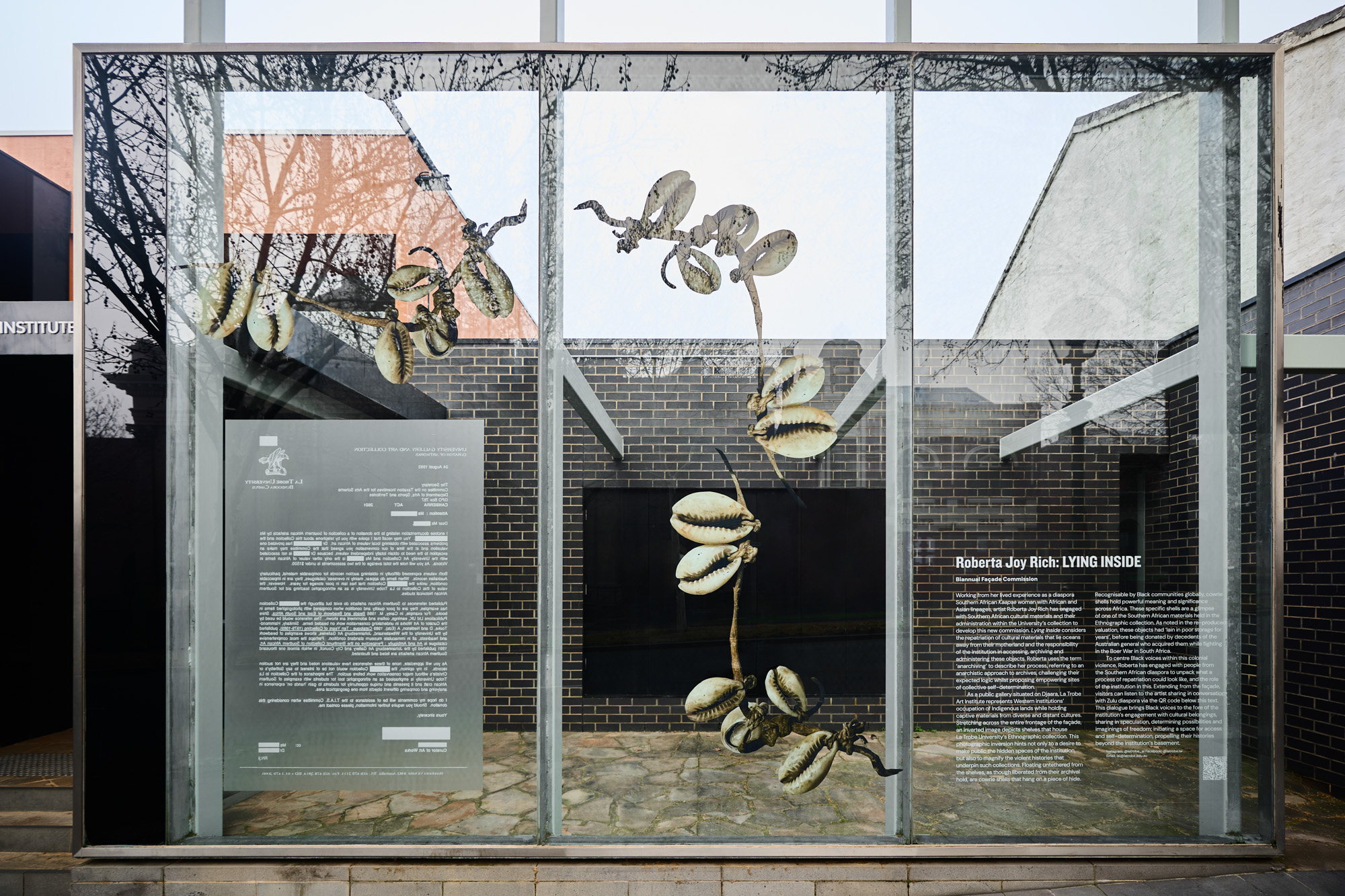 Glass display vinyl prints with cowrie shell clusters, a mirrored redacted university letter, and exhibition text about the artwork.
