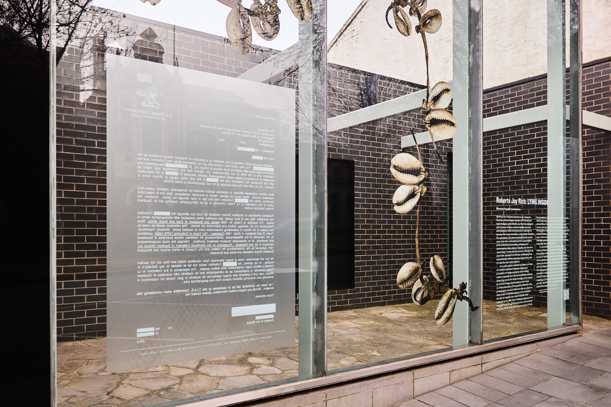 Close view of a mirrored, partly redacted 1993 university letter printed on glass, with cowrie shells on a hide strand at right.