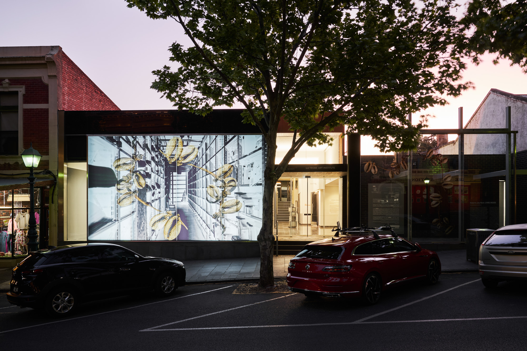 The lit window artwork glowing at dusk, with parked cars in front and the gallery foyer and stairs visible through the entrance doors.
