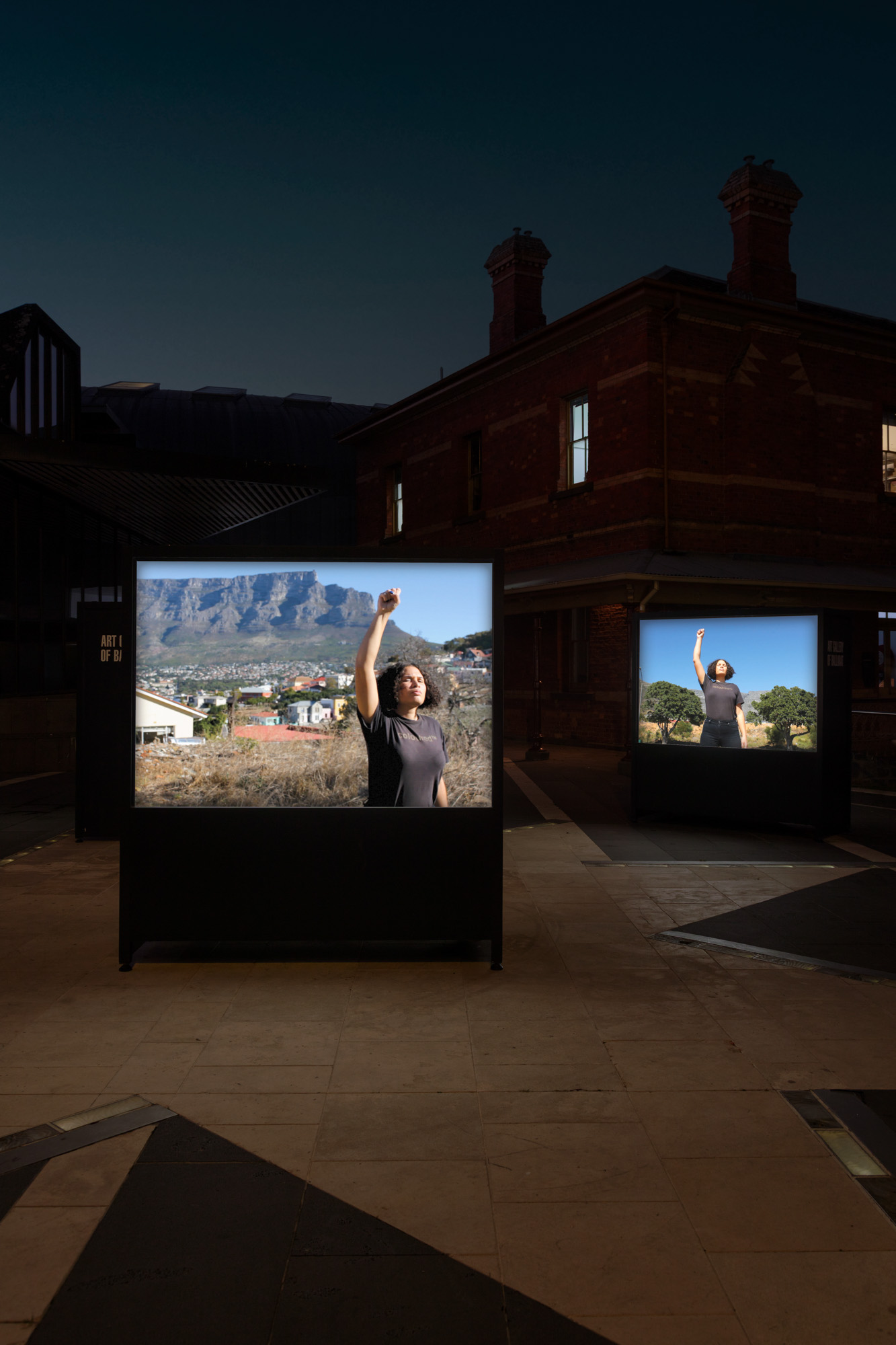 Two glowing screens in a dark brick courtyard show a Black woman raising her fist against views of Table Mountain, Cape Town, and trees lining a road in District Six.