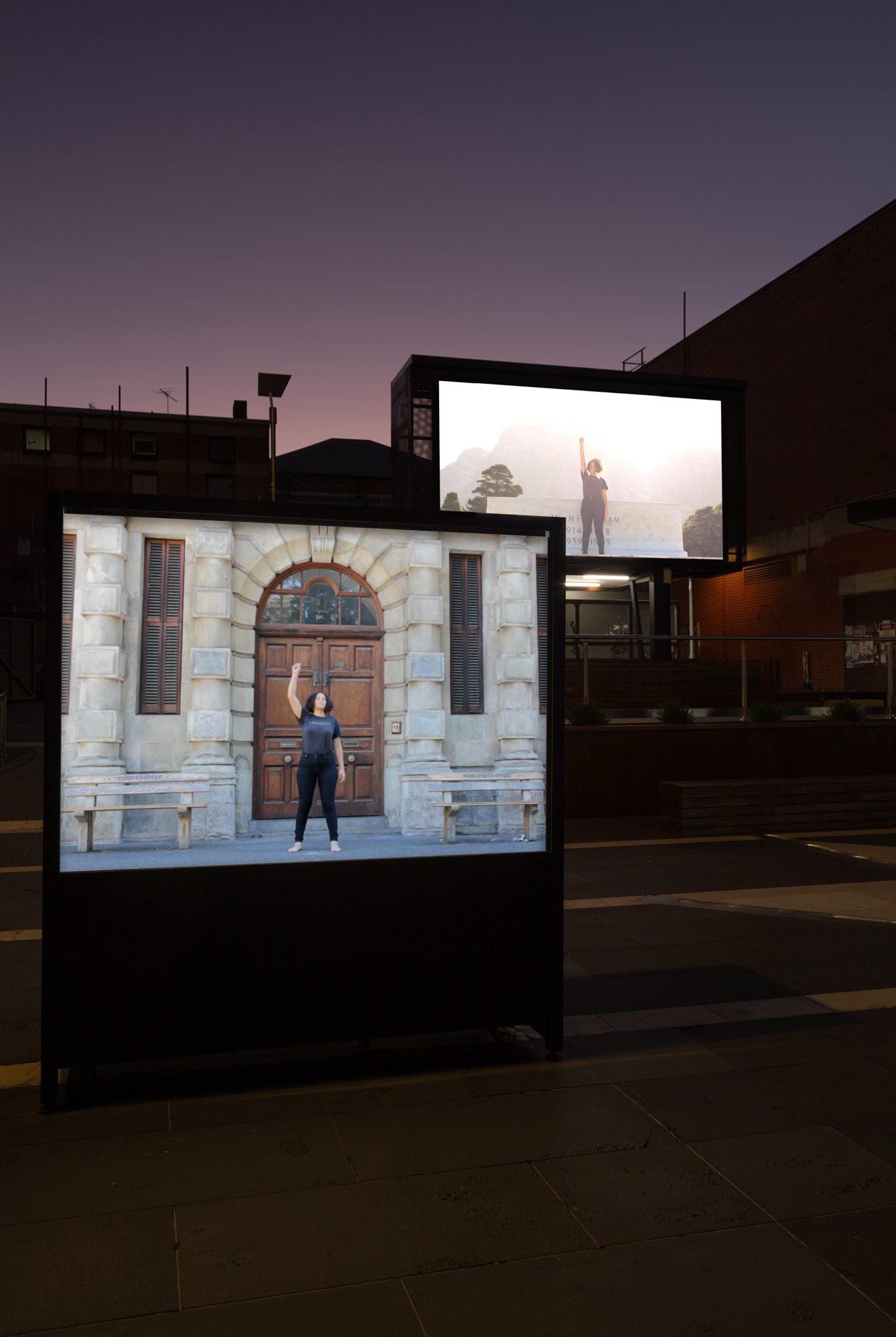 Two illuminated outdoor screens at dusk show a Black woman raising her fist—one outside an ornate wooden doorway and the other against a mountain landscape.