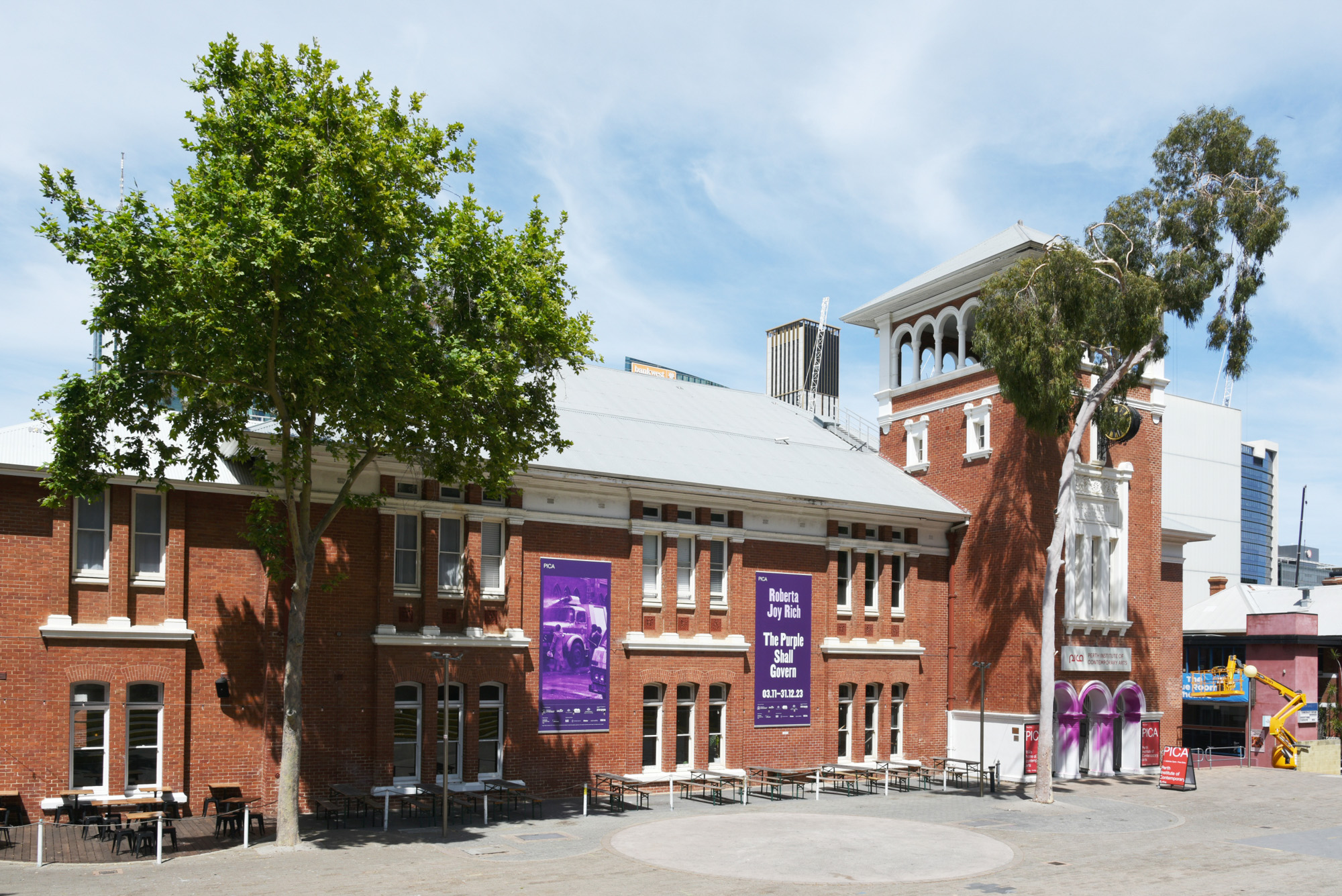 Wide daytime view of the Perth Institute of Contemporary Arts, a red-brick heritage building with purple exhibition banners, framed by tall trees, purple-painted entrance arches and a paved courtyard.