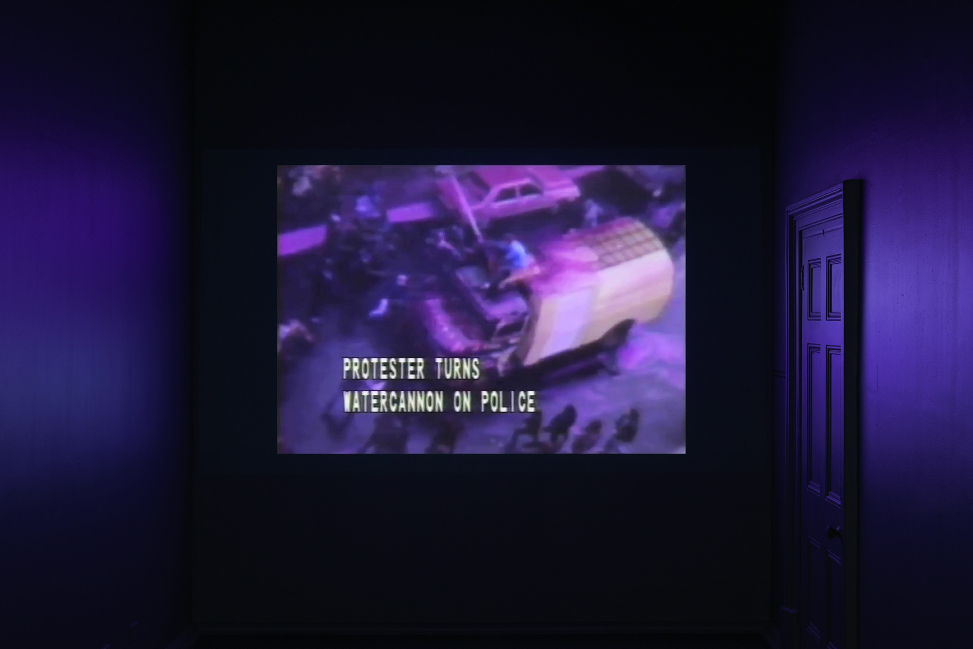 A dark gallery bathed in purple light displays archival protest footage of a water cannon, captioned “Protester turns watercannon on police.”
