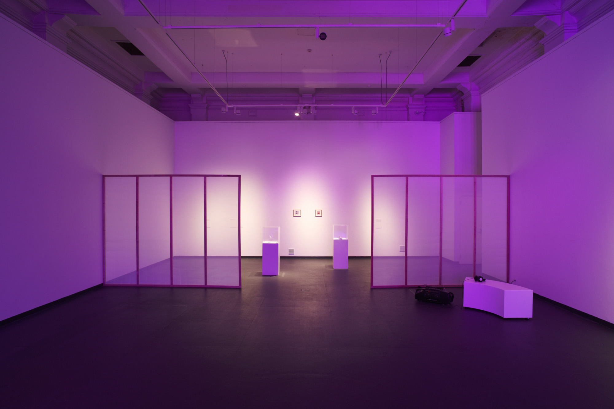 Wide view of a purple-lit gallery with two translucent partitions framing display cases and small photographs, with audio equipment and seating to the right.