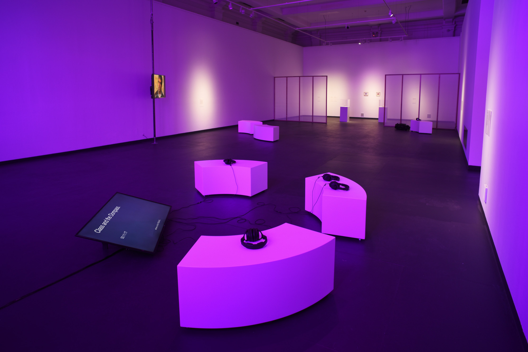 Wide view of a purple-lit gallery with curved white listening seats, headphones, a floor-level video screen, suspended screen on a metal pole playing a video work, display cases, and translucent partitions.