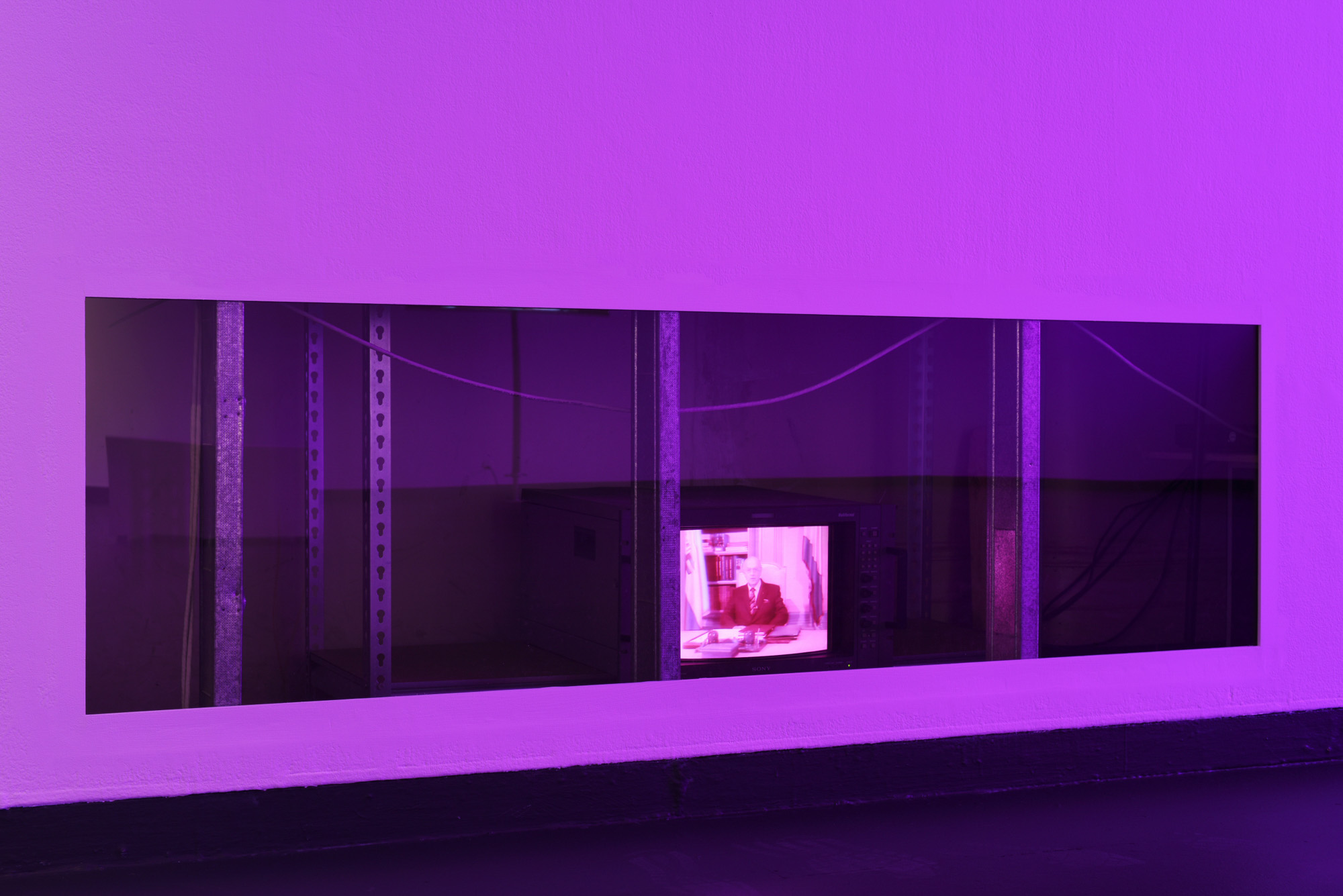 A small vintage broadcast monitor playing archival footage of South African politician P.W. Botha seated at a desk is visible through a long, dark opening cut through the base of a purple-lit gallery wall.