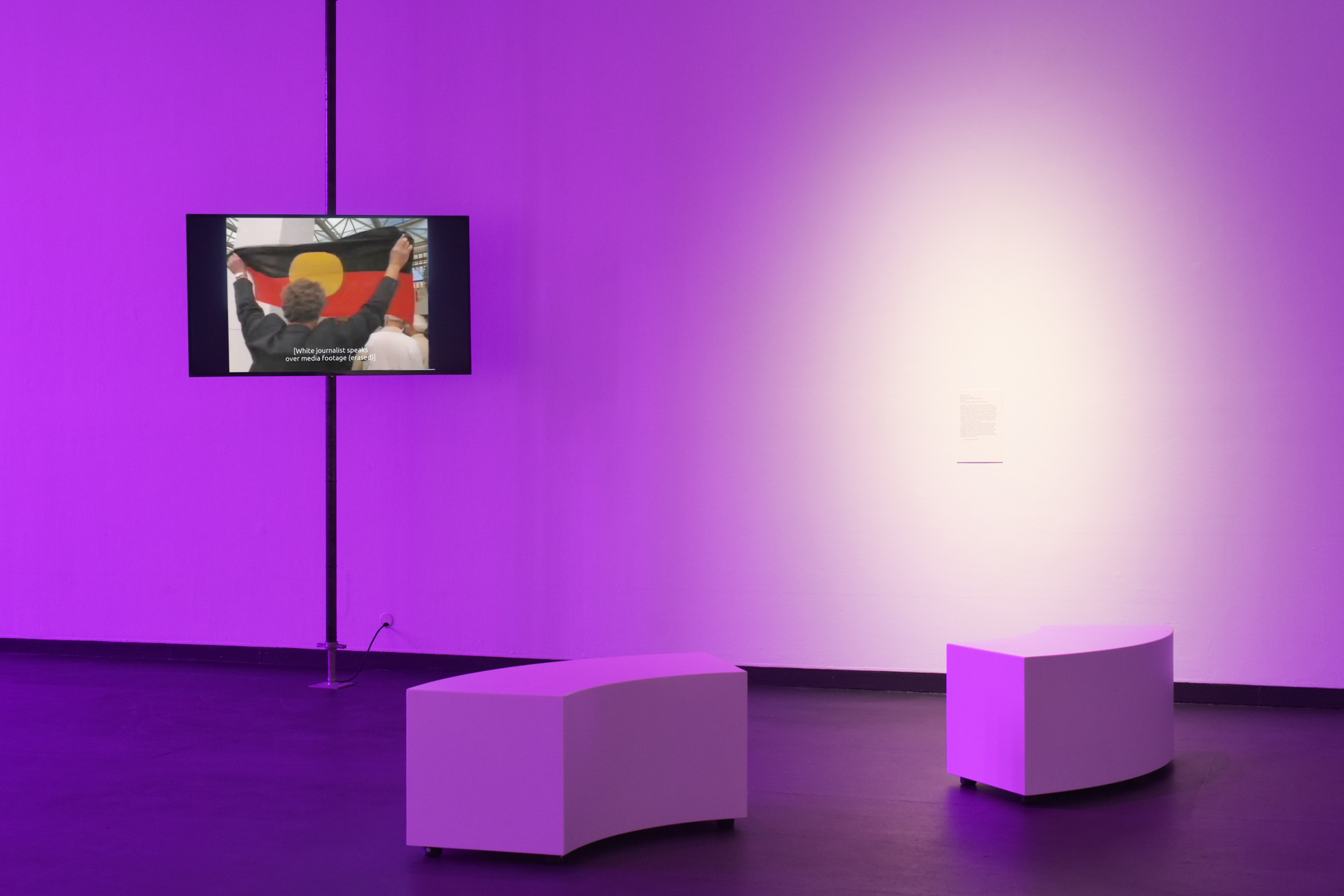 Suspended screen shows an Indigenous person holding up the Aboriginal flag, with two curved white benches positioned in the purple-lit gallery. A white spotlight circles the gallery wall label.