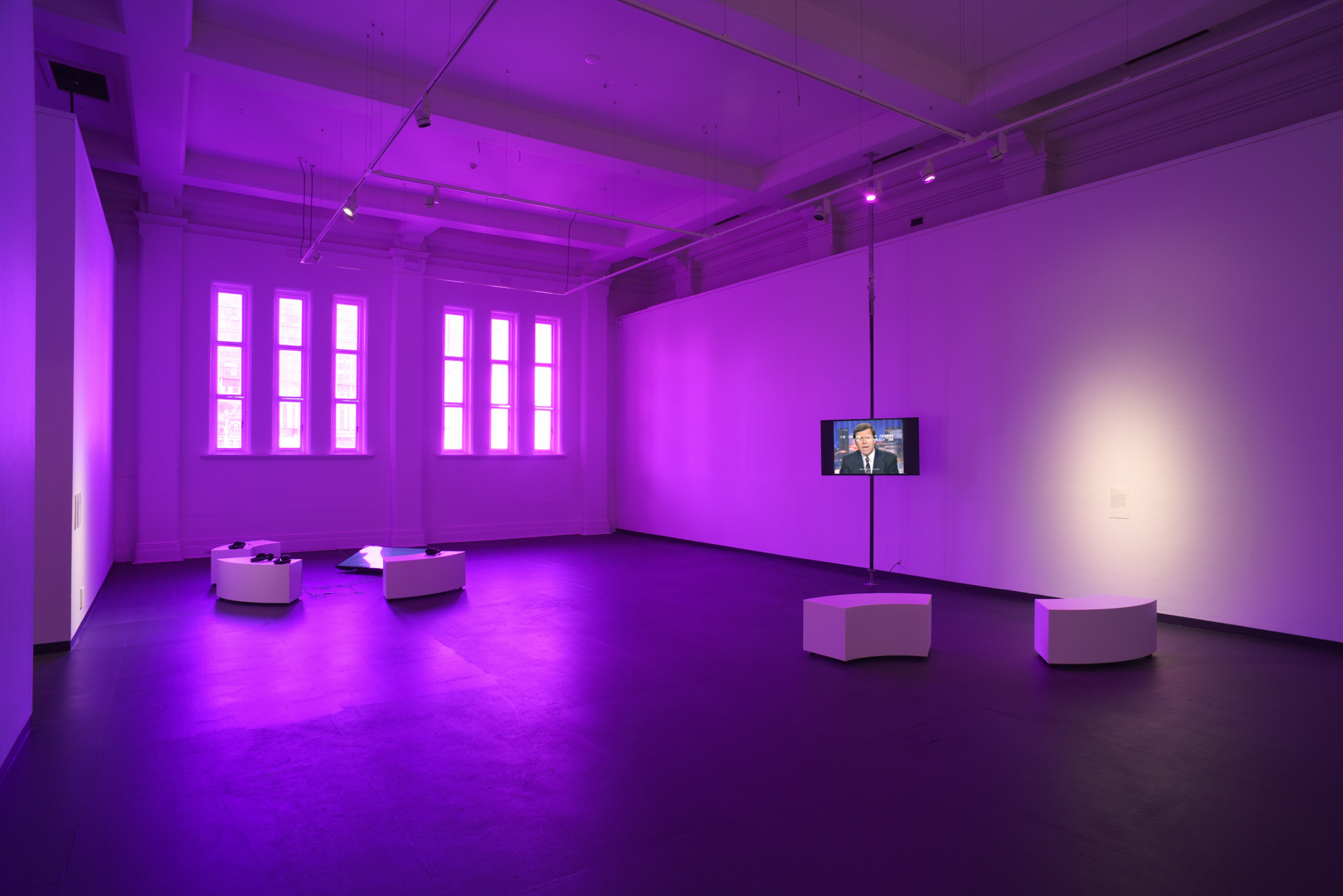 Wide view of a spacious purple-lit gallery with tall windows, curved white listening benches, headphones, and a suspended screen on a metal pole plays an archival video of 90s news media showing a man in a suit.