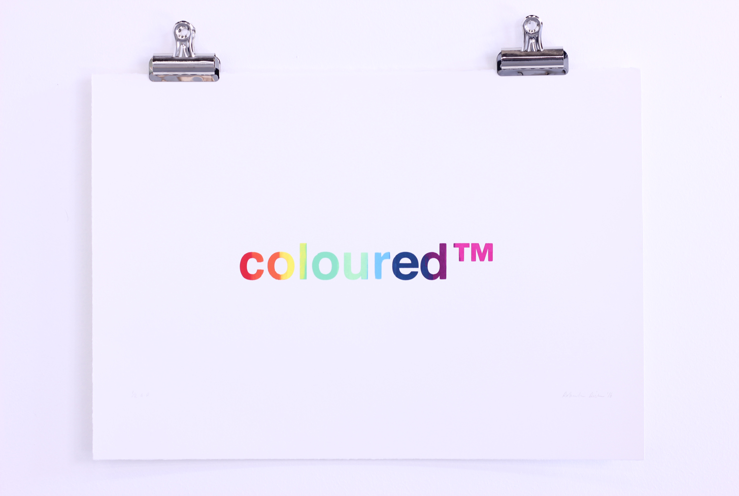 A serigraph print on white paper held by two silver clips display the word “coloured™” in rainbow-coloured lowercase letters, against a white wall.