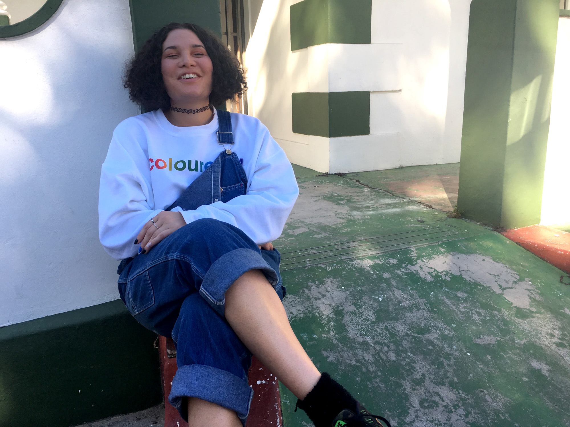 A smiling young light skinned Black woman in denim overalls and a white “coloured™” sweatshirt sits cross-legged outside a white-and-green building.