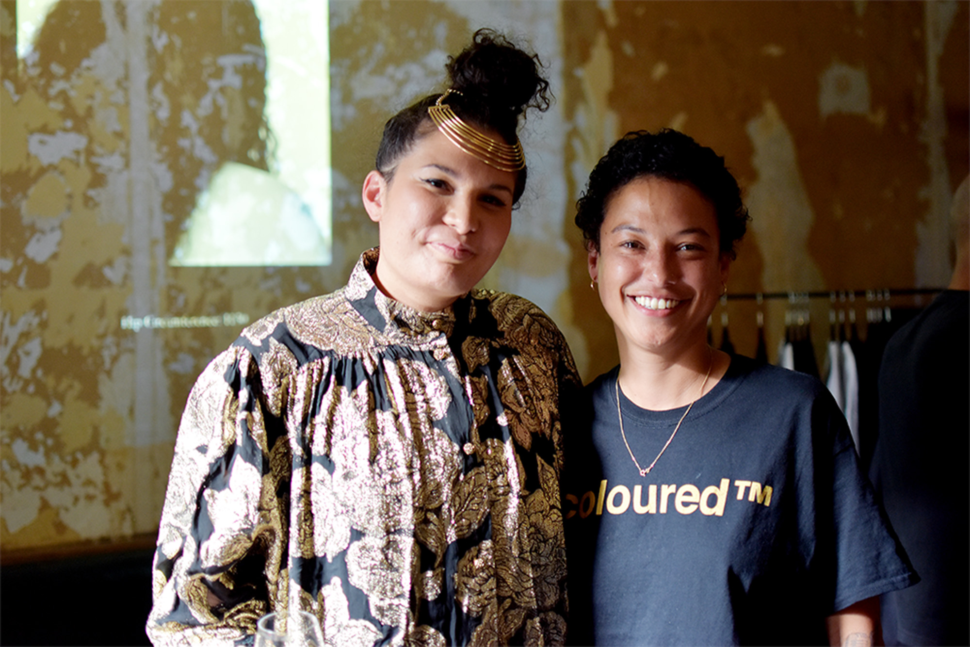 Two light skinned Black South African women are smiling and standing together indoors; one wears an ornate gold-and-black floral top and gold headpiece, while the other wears a black “coloured™” T-shirt with gold lettering.