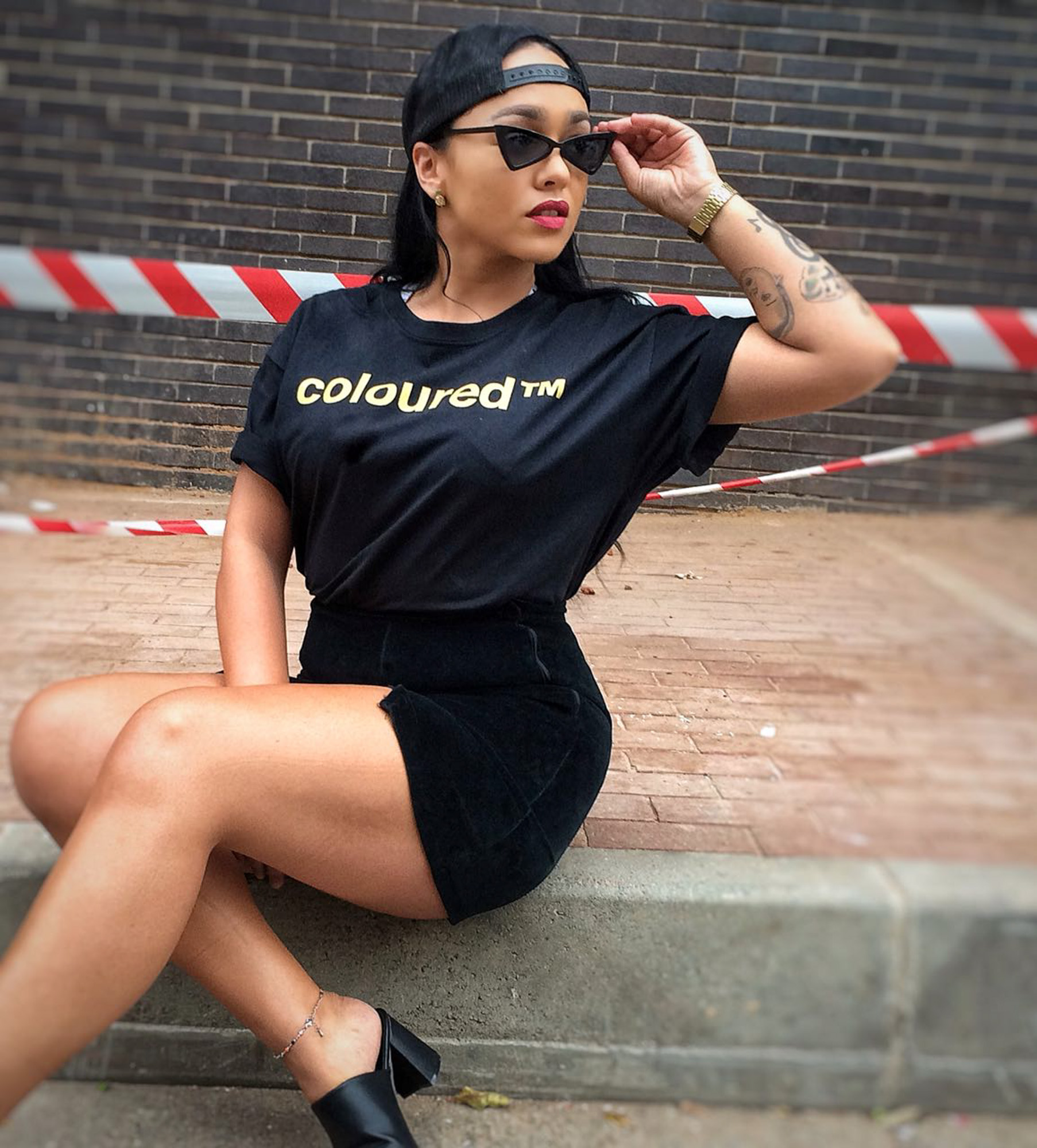 A light skinned Black South African woman wearing sunglasses, a backward cap, black shorts, and a black “coloured™” T-shirt poses on a low wall, with red-and-white barrier tape behind them.