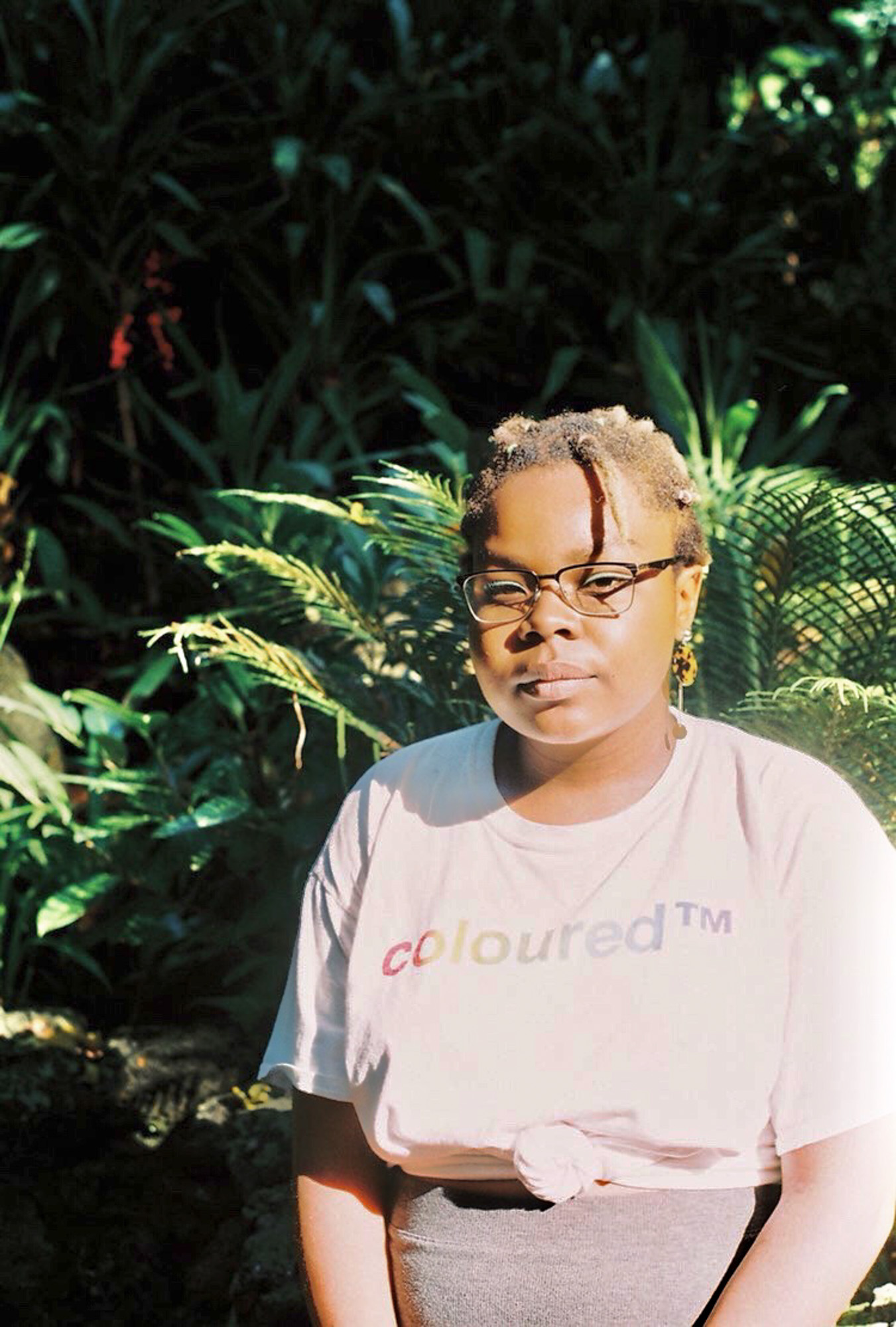 A young Black woman with short natural hair, pale green eyeshadow and glasses sits in bright sunlight among dense tropical plants, wearing a white “coloured™” T-shirt with rainbow lettering.