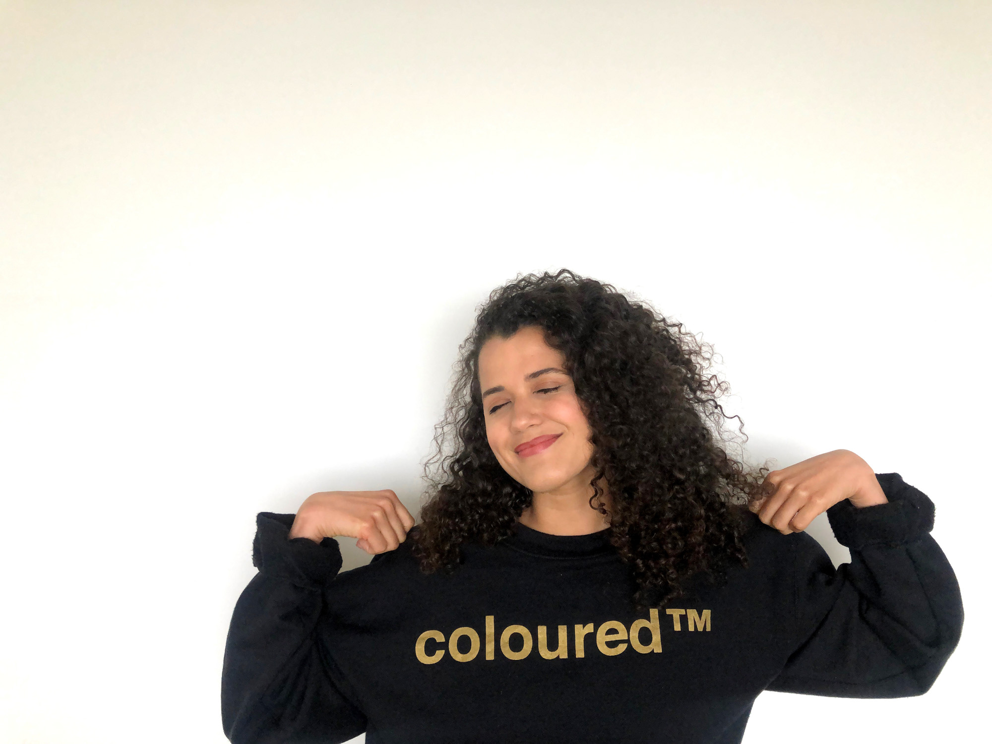 A smiling light skinned Black South African woman with long curly hair holds up their black sweatshirt with gold “coloured™” lettering while standing against a plain white wall.
