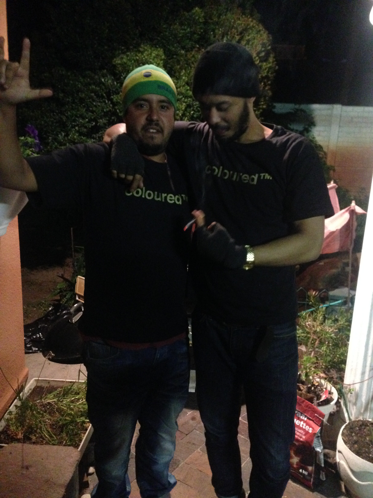 Two Black South African men are wearing black “coloured™” T-shirts stand with their arms around each other in a garden at night; one raises a hand toward the camera.