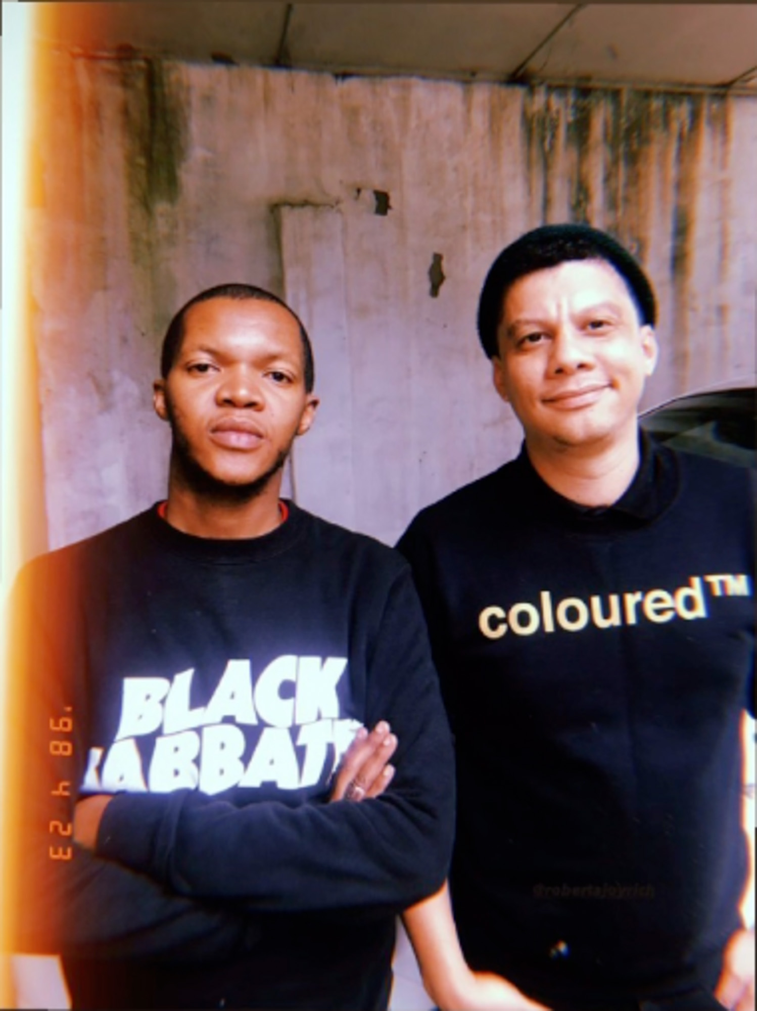 Two Black South Africans, one light skinned, pose in front of a weathered concrete wall; one wears a black “Black Sabbath” sweatshirt and the other a black “coloured™” T-shirt with gold lettering.