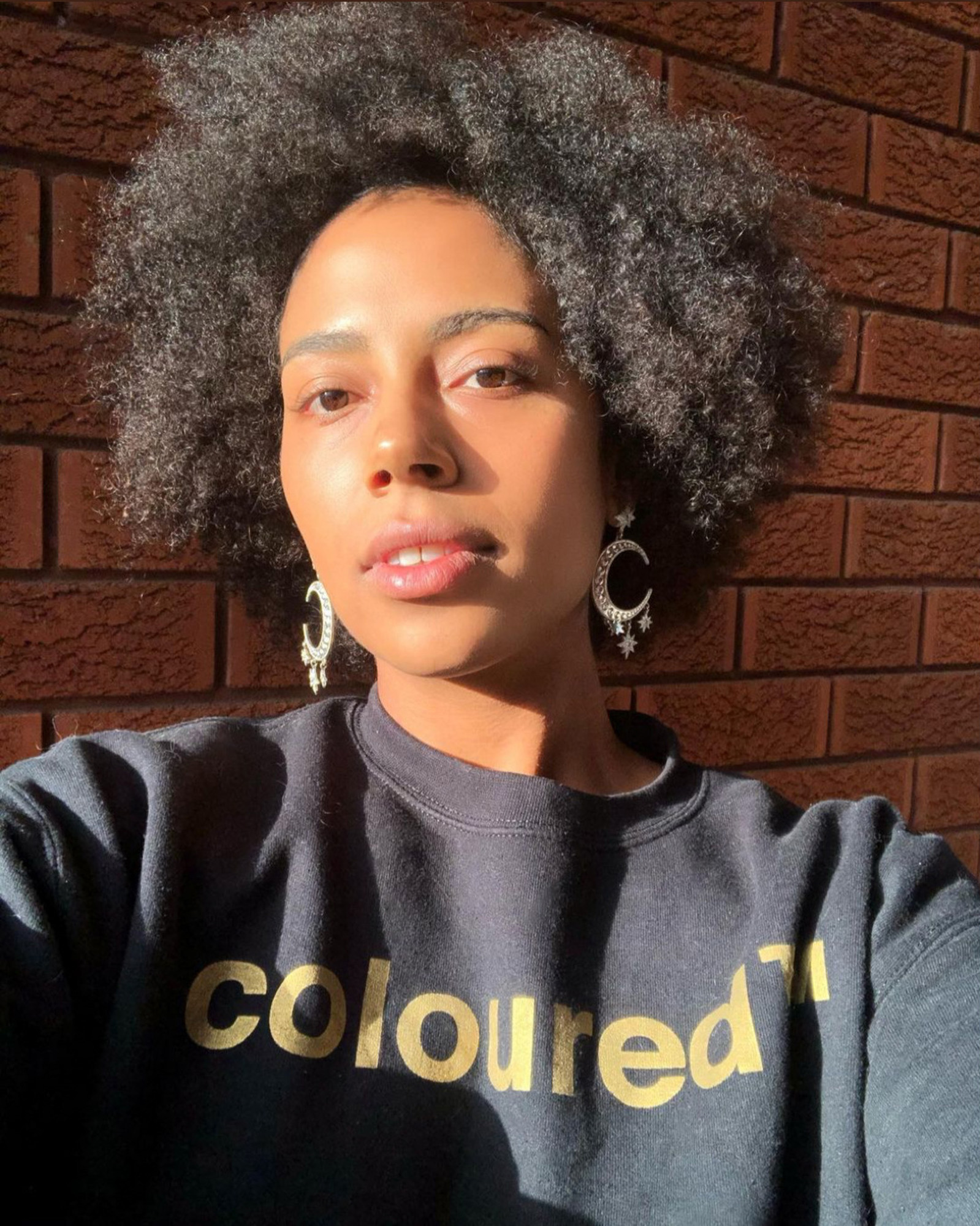 Sunlit close-up selfie of Black Tūhoe Grenadian woman with natural hair and crescent moon earrings wearing a black “coloured™” sweatshirt against a red brick wall.