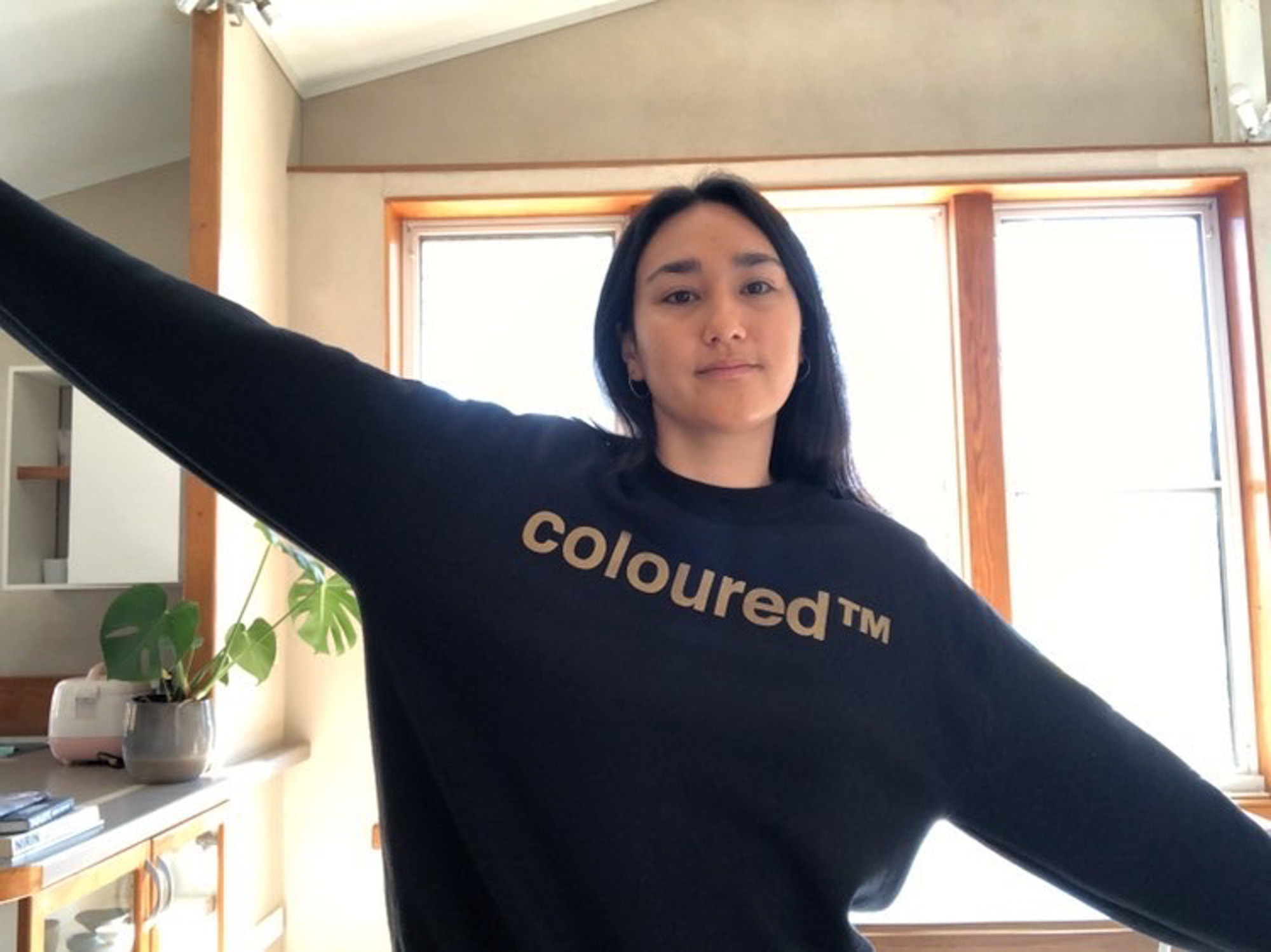 A diaspora woman with Korean heritage is wearing a black “coloured™” sweatshirt extending both arms outward in a bright room with large windows and indoor plants.