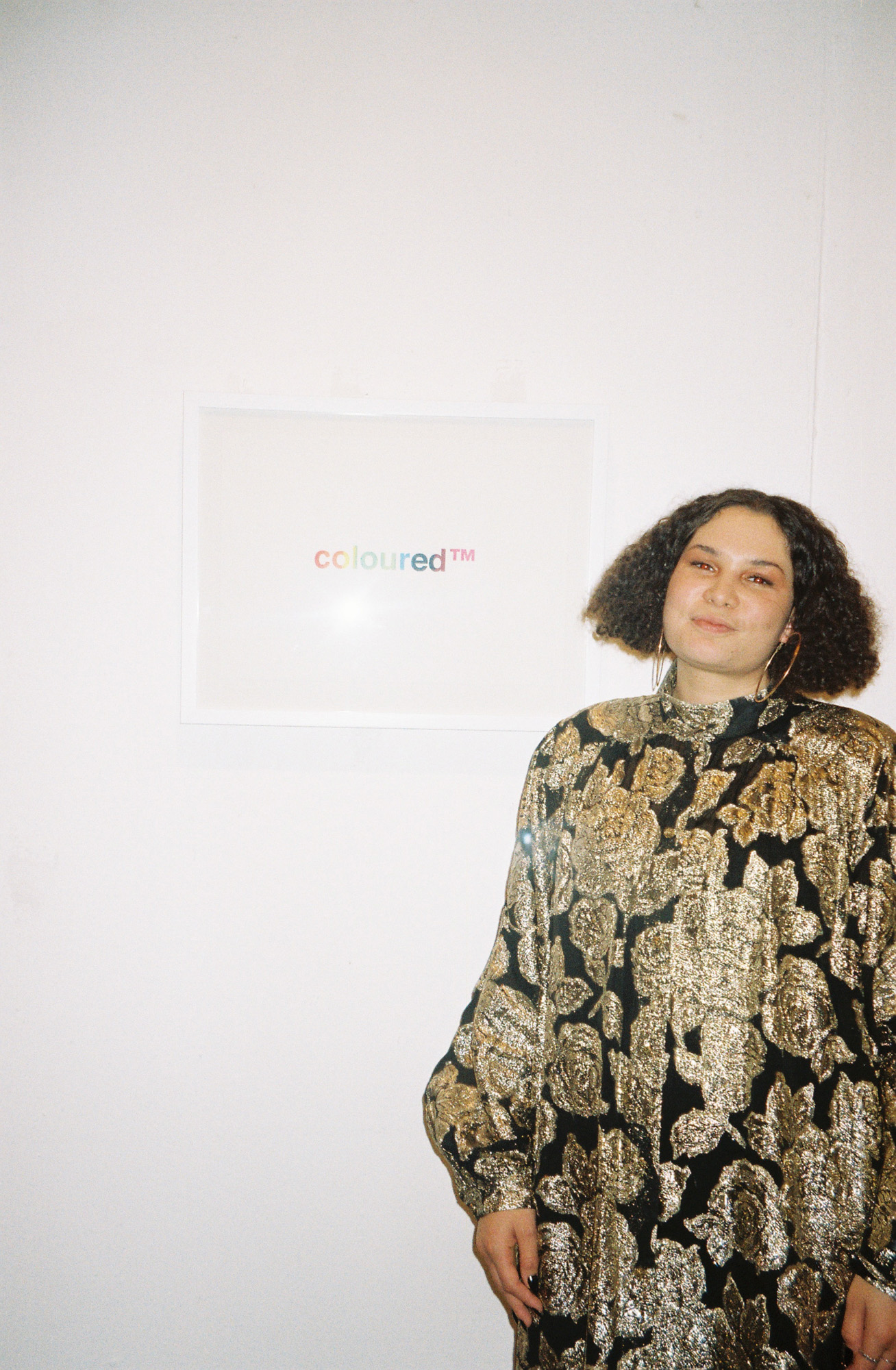 A light skinned Black South African woman wearing a metallic gold-and-black floral outfit stands beside a framed print displaying “coloured™” in rainbow letters on a white gallery wall.
