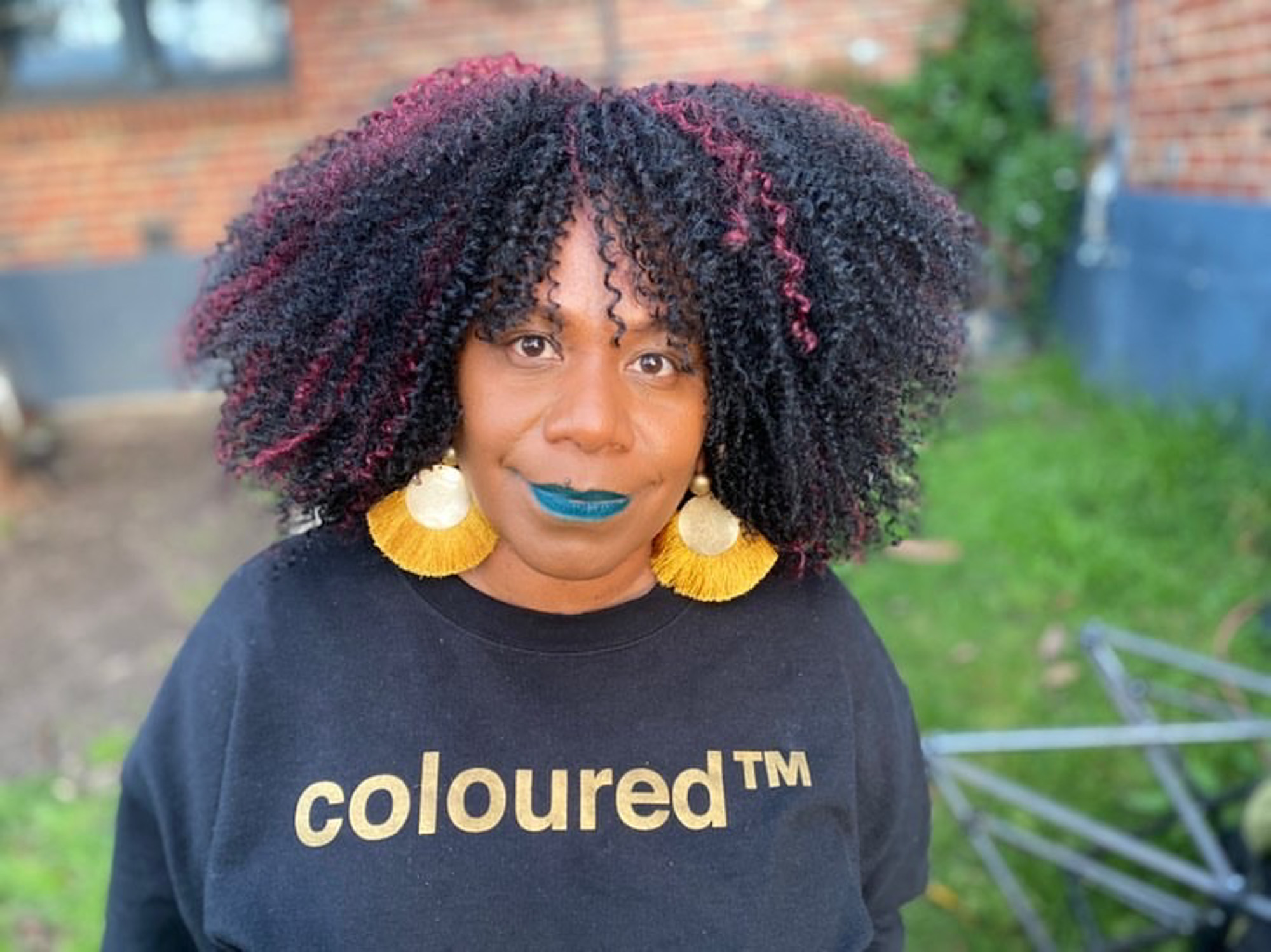 A Black Papua New Guinea Tolai woman with voluminous black-and-pink curls, teal lipstick, and large gold tassel earrings looks at the camera while wearing a black “coloured™” sweatshirt outdoors.