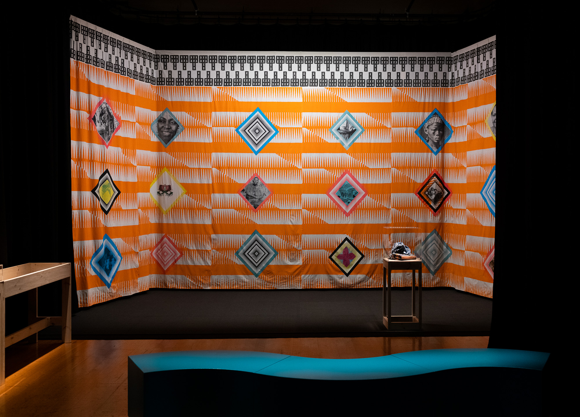 A dark gallery displays a large, colourful, three-sided textile installation with a repeated orange-and-white striped pattern, with black-and-white borders, and diamond-framed portraits with geometric designs and Black women icons that hangs from the ceiling. A small display table and curved teal bench sit in front adjacent to a vitrine displaying African fabrics folded inside.