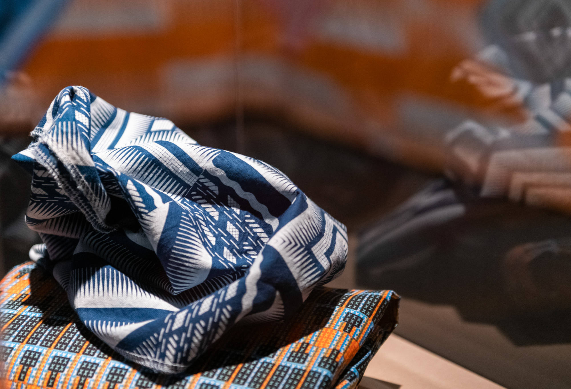 Close-up of shweshwe fabric of blue-and-white bold geometric and organic patterns rests on an orange, teal, and black folded African textile, within a perspex display case reflect the fabrics upon its surface as well as Aunties Curtain blurred in the background.