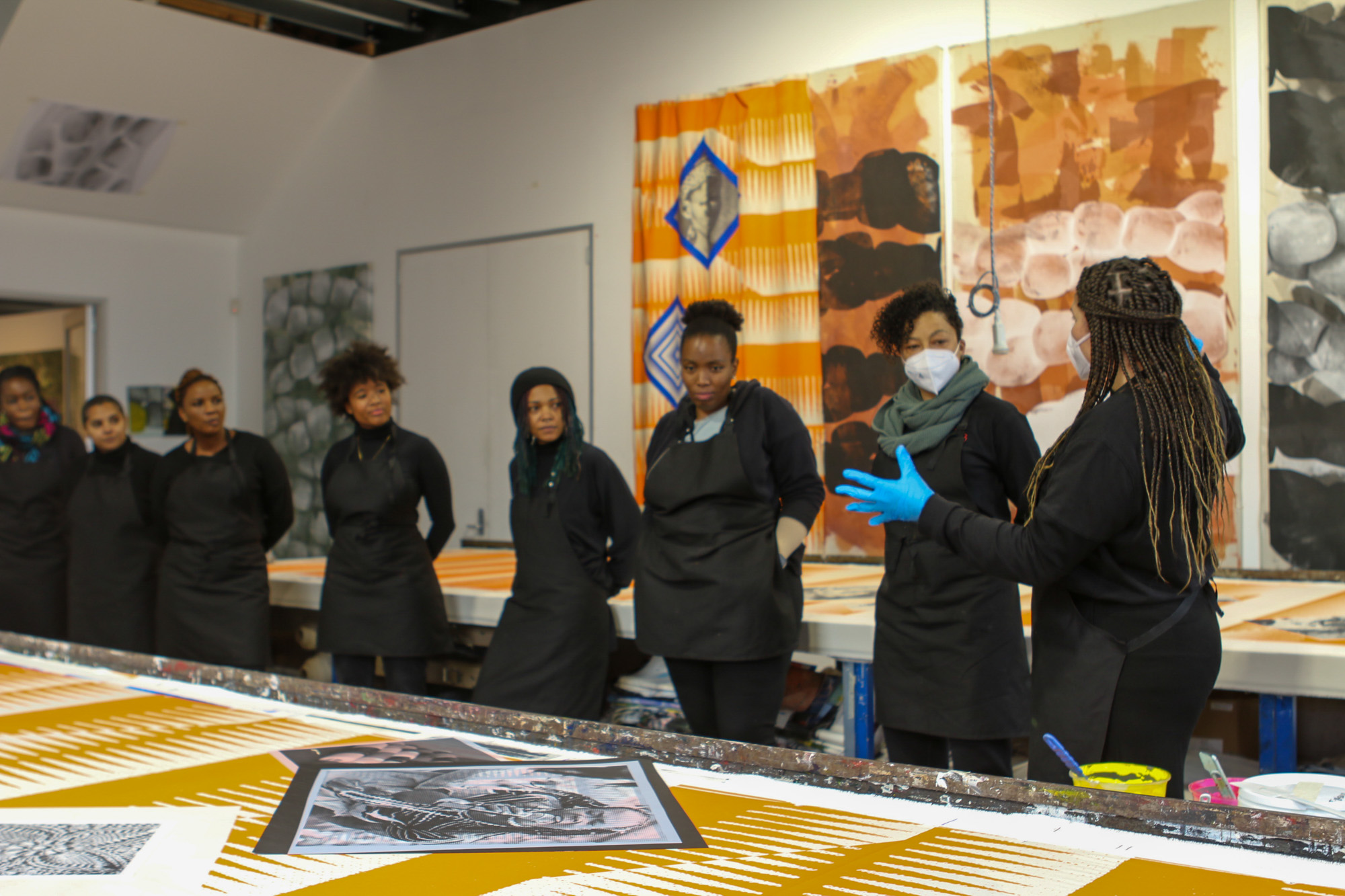 A group of eight Black women artists dressed in black clothing and aprons gather around screen-printing tables in a bright studio. An artist with long braids and blue gloves addresses the group, surrounded by large orange, brown, black, and white patterned textiles.