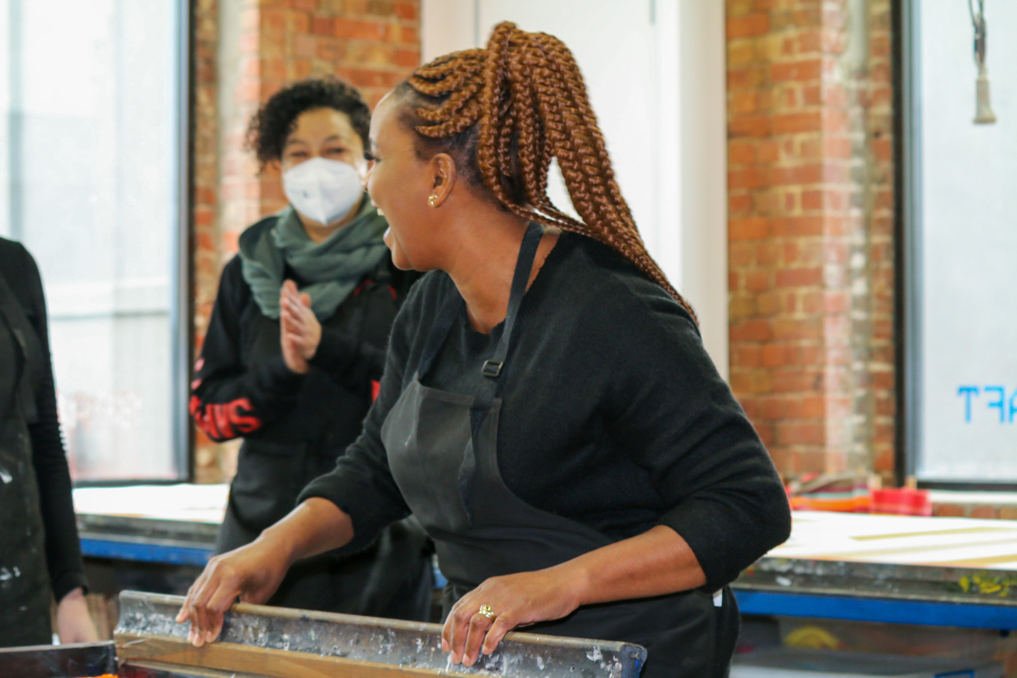 A woman artist with long copper-coloured braids pulls a wooden squeegee across a large screen-printing frame. A masked colleague stands behind her with hands raised clapping, against a backdrop of tall windows and exposed brick walls.