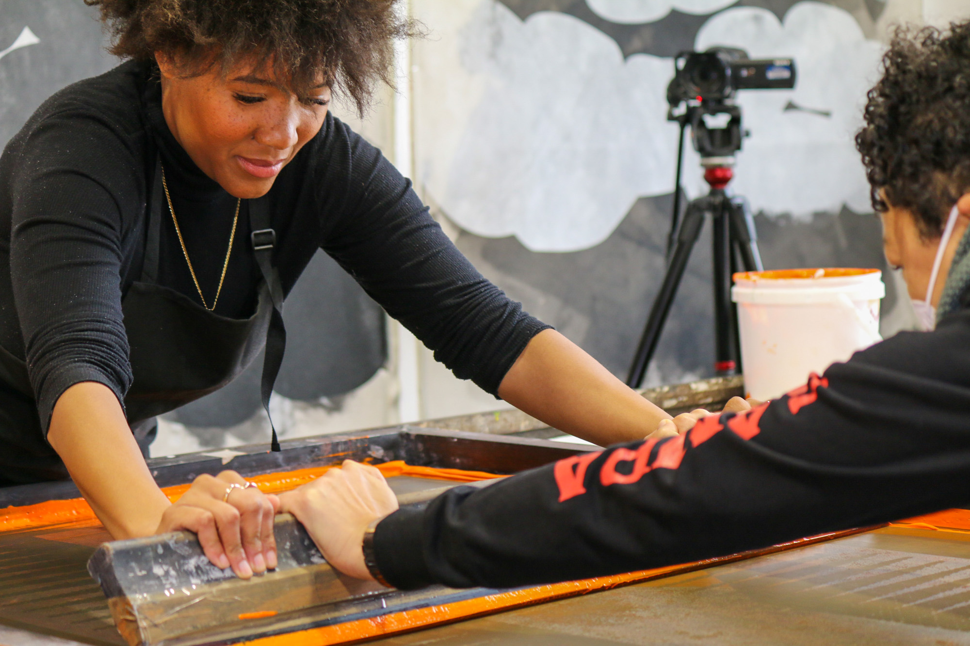 Two Black women artists work together to pull a large metal squeegee across a screen filled with vivid orange ink. A video camera on a tripod records the printing process against a backdrop of black-and-white painted textile forms.