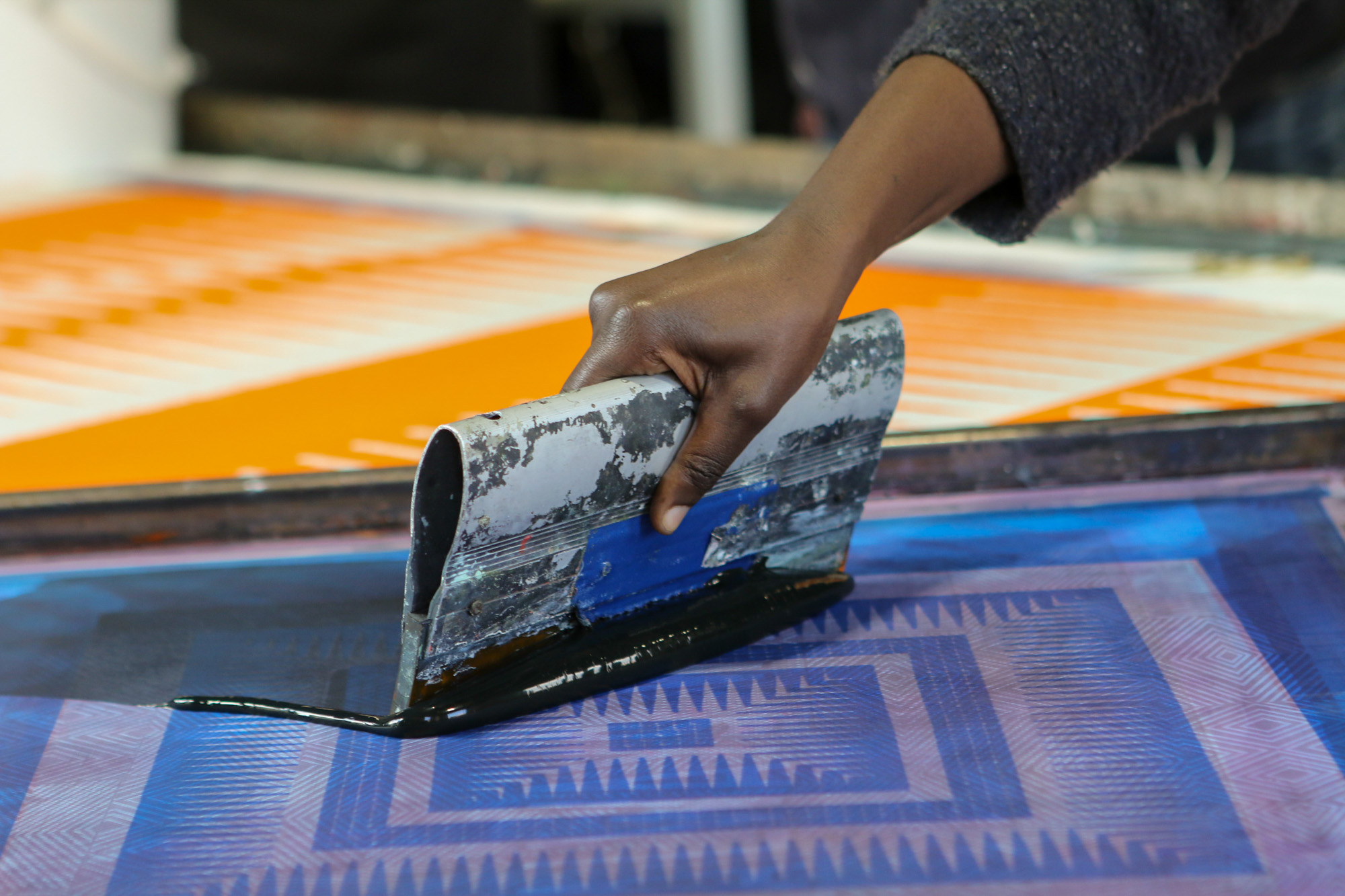 Close-up of a Black woman artist’s hand gripping a worn metal squeegee and pulling black ink across a screen containing a blue and purple geometric design. A completed orange-and-white print lies blurred on the table behind.