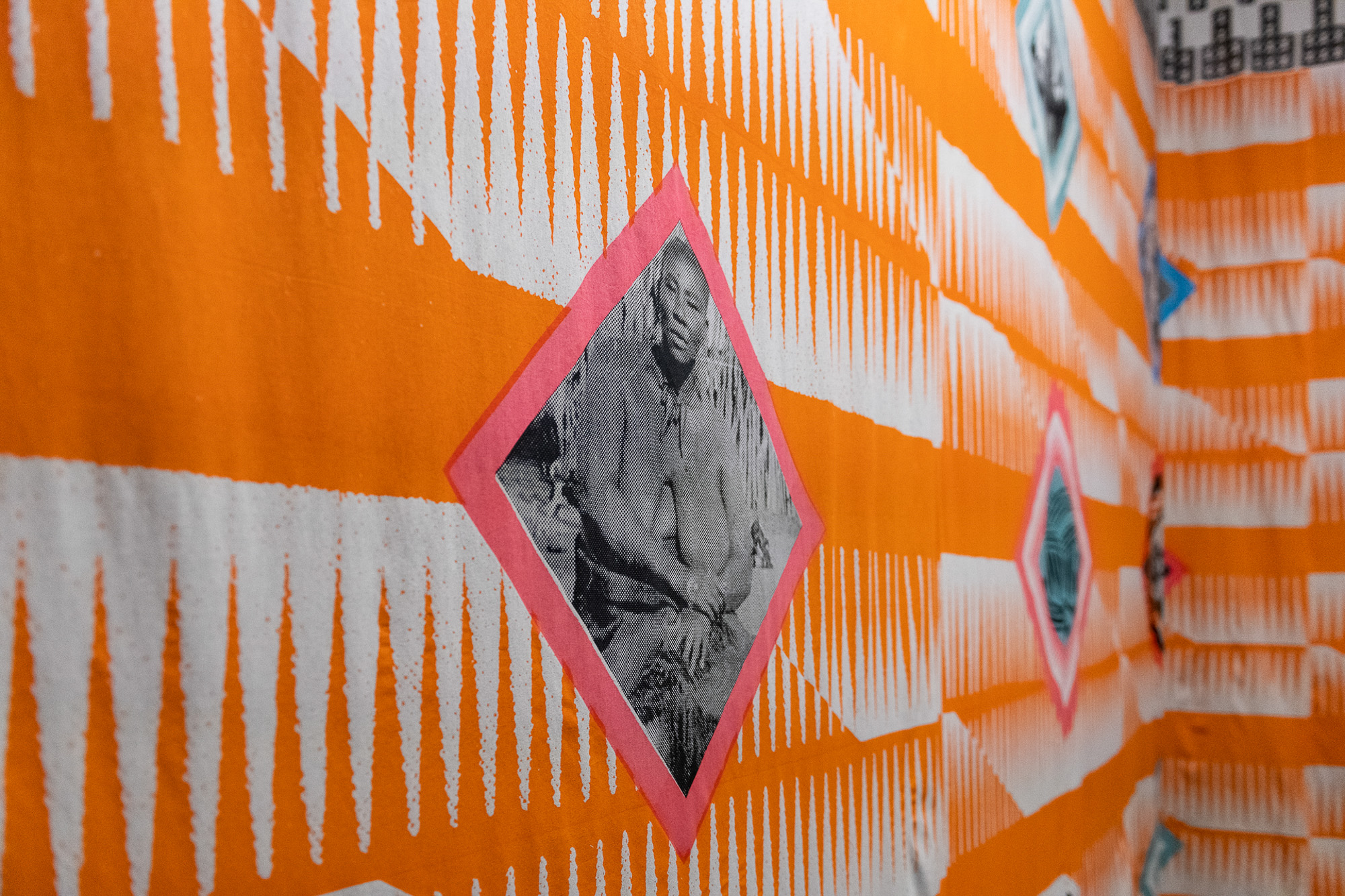 Close-up of a pink diamond with a black-and-white print of a seated Black woman, is part of the large screen printed textile installation with bright orange-and-white repeated comb-like linear patterns.