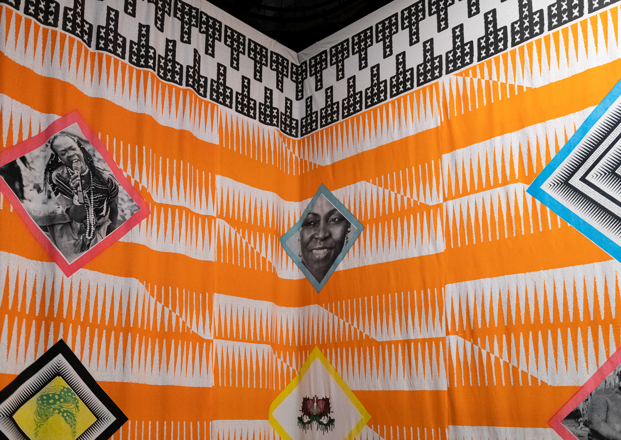 Detailed corner view of a large colourful textile installation, showing fabric covered in an orange and white repeating comb-like stripe pattern and topped with a black-and-white geometric border. Diamond-framed portraits of Black women and abstract designs in pink, blue, yellow, and black are arranged across the fabric.