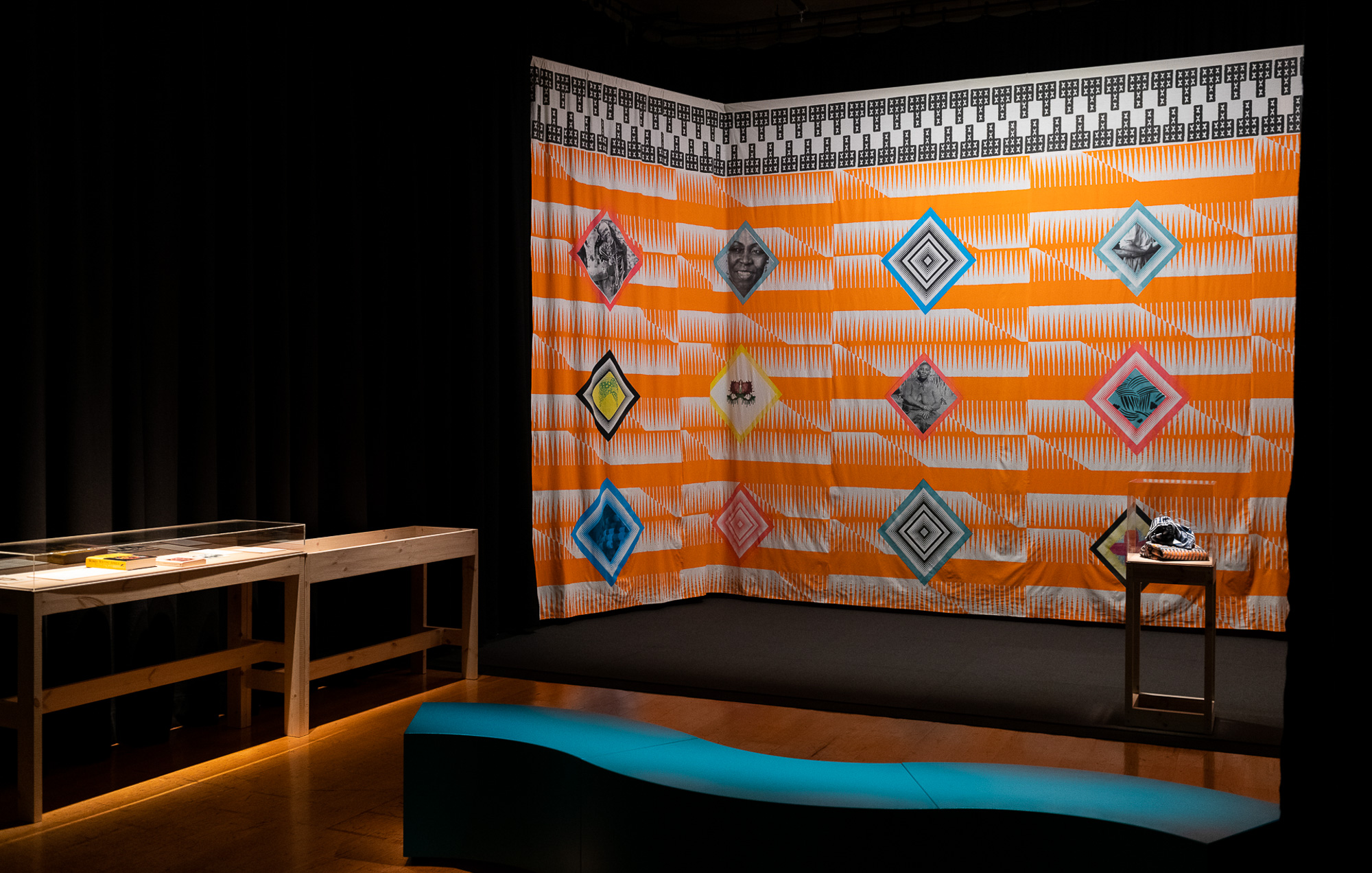Wide view of the darkened gallery, where the large orange-and-white patterned textile installation is decorated with colourful diamond-framed black-and-white photographs of Black women and geometric motifs. Wooden display cases, a small pedestal displaying folded fabric, and a curved teal bench occupy the foreground.
