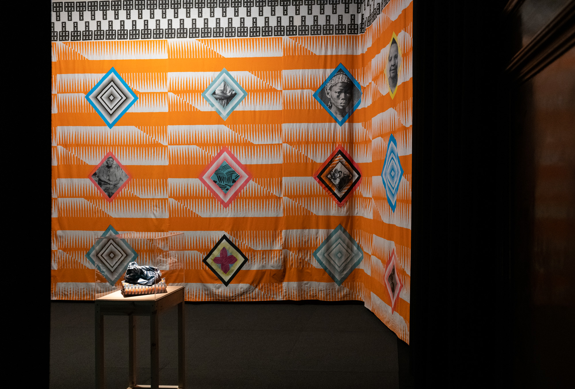 Side view into the darkened gallery of the large colourful installation, showing the right-hand section of the orange-and-white comb-like patterned textile. Diamond-framed portraits of Black women and abstract designs cover both walls, while a small wooden pedestal displays blue-and-white shweshwe fabric resting upon folded orange-white-black-teal African fabric in front.