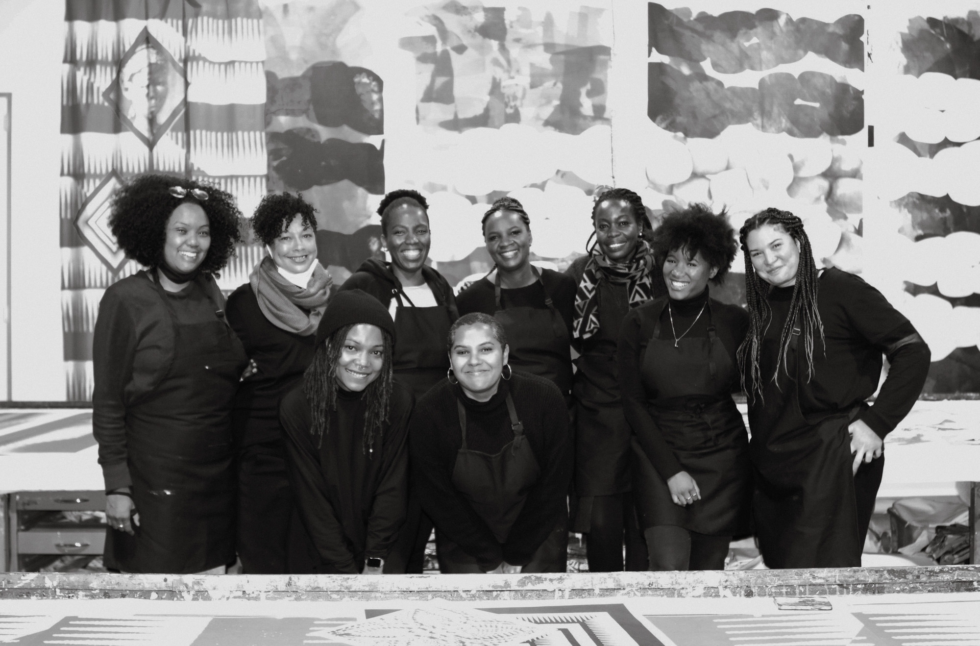 Black-and-white group portrait of nine smiling African women artists wearing black clothing and aprons in a screen-printing studio. Seven stand behind two crouching colleagues, surrounded by printing tables and large patterned artworks.
