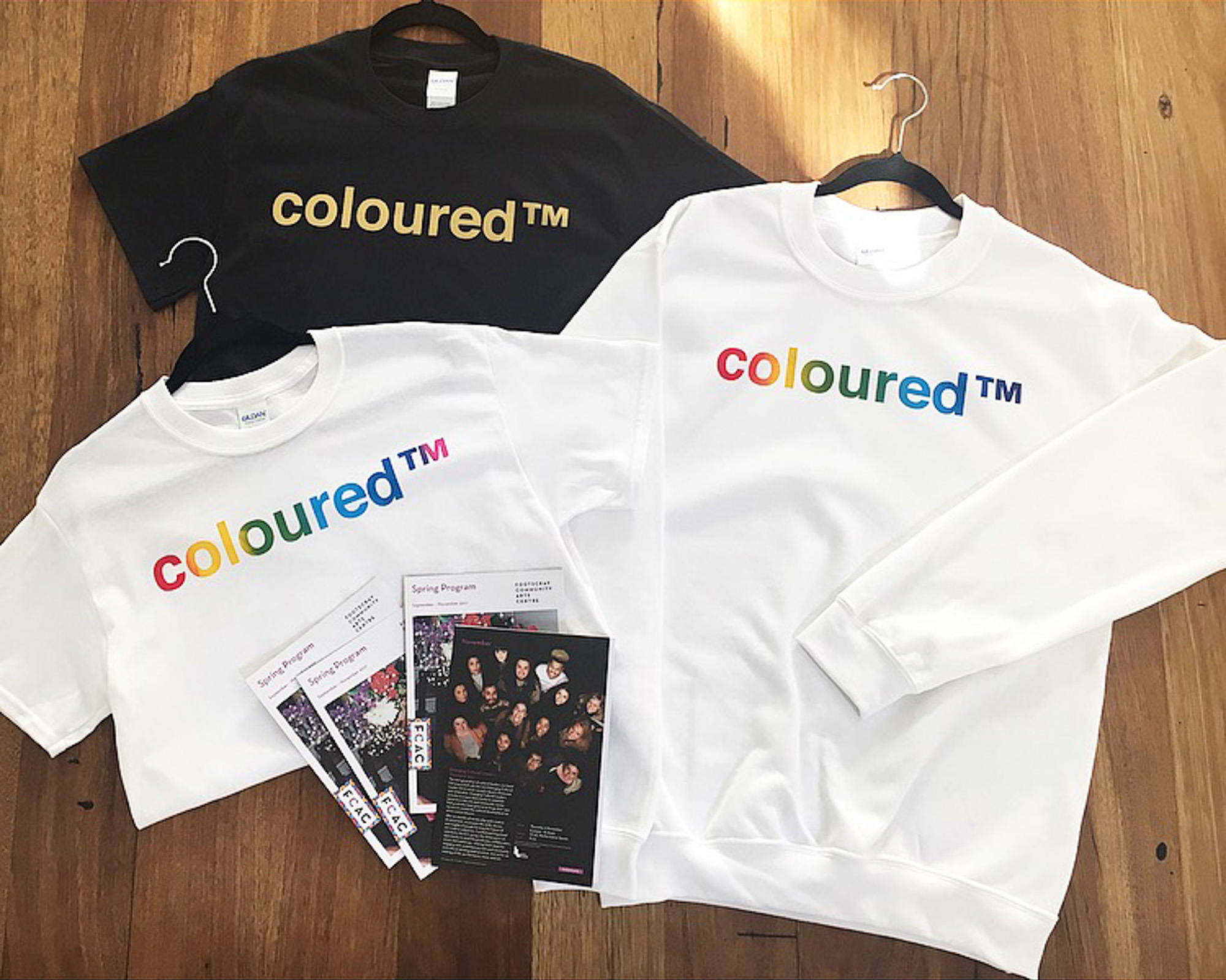 Black and white T-shirts and a white sweatshirt printed with “coloured™” in gold on black, and rainbow lettering on white, arranged with Footscray Community Arts programs on a wooden table.