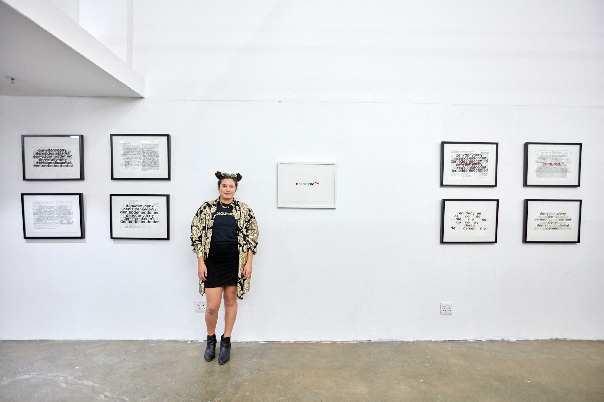 Artist Roberta Joy Rich stands against a white gallery wall between framed text-based prints. Eight black-framed works repeat the words "deny, denial, denied" over archival documents, and a white-framed print reads "coloured™" in rainbow letters.