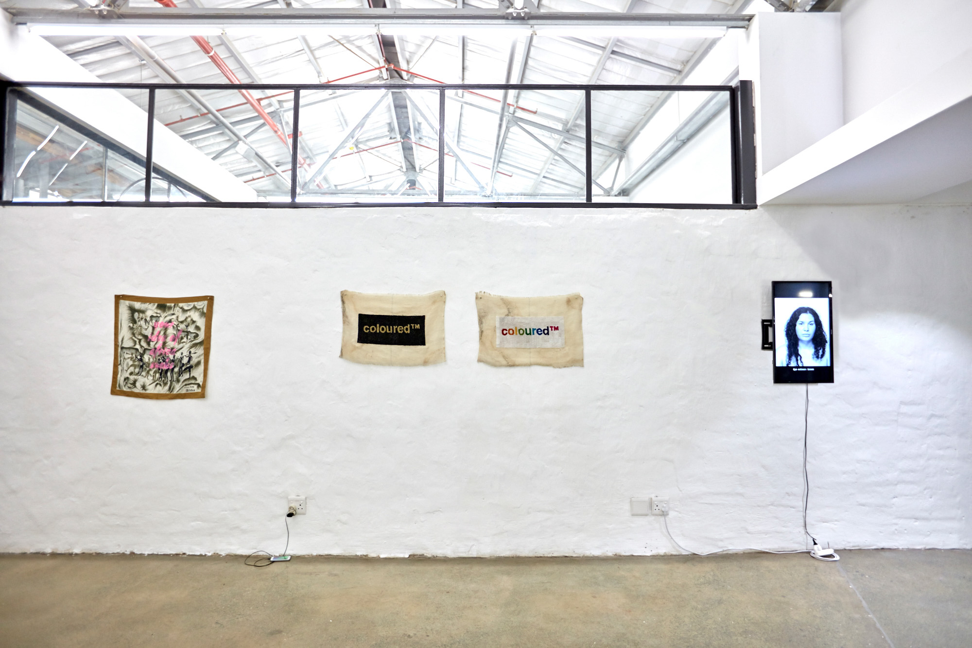 White brick gallery wall beneath an industrial glass ceiling. Displayed are a vintage Africa-themed scarf overpainted with pink text, two frayed tapestries reading "coloured™" — one gold on black, one rainbow on white — and a wall-mounted screen showing a black-and-white portrait of a woman.