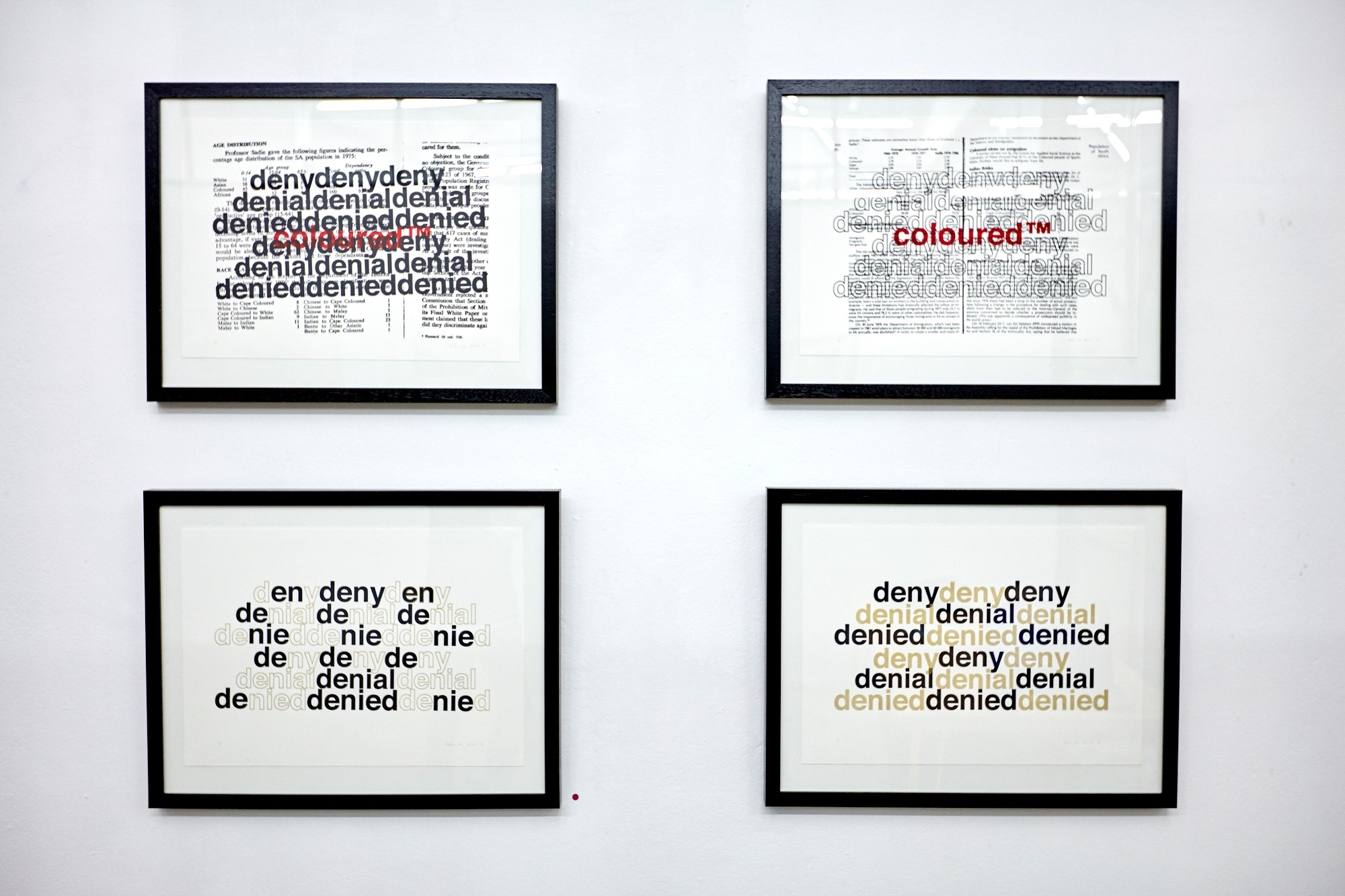 Four black-framed prints in a grid. The top two overlay apartheid-era documents with the repeated words "deny, denial, denied" and "coloured™" in red; the bottom two repeat the same words in black and gold type, one interweaving the Afrikaans "nie."