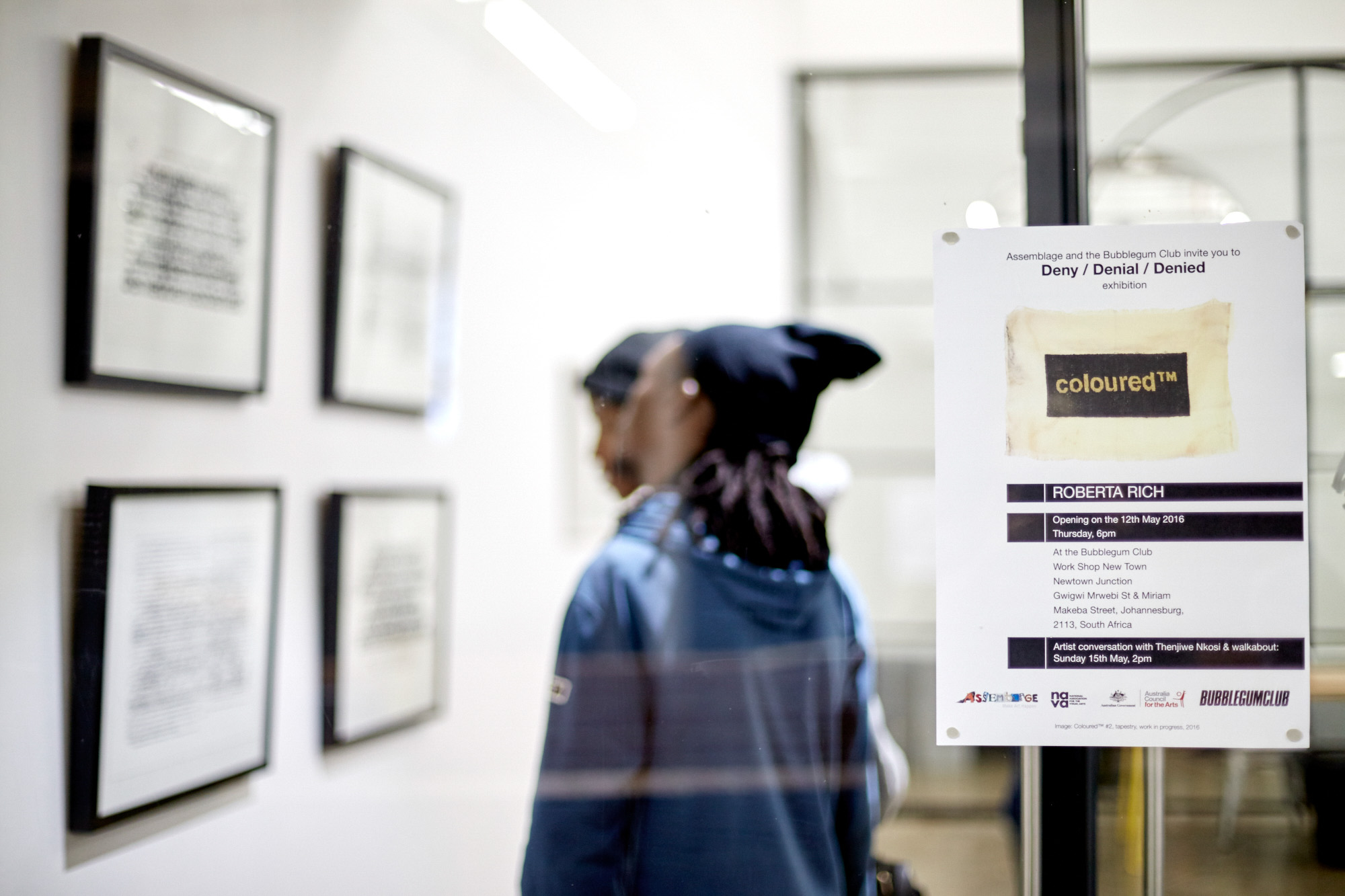 A poster on a glass door invites visitors to the Deny / Denial / Denied exhibition by Roberta Rich, opening 12 May 2016 at the Bubblegum Club, Johannesburg, with an artist conversation with Thenjiwe Nkosi. A visitor and framed prints are visible through the glass.