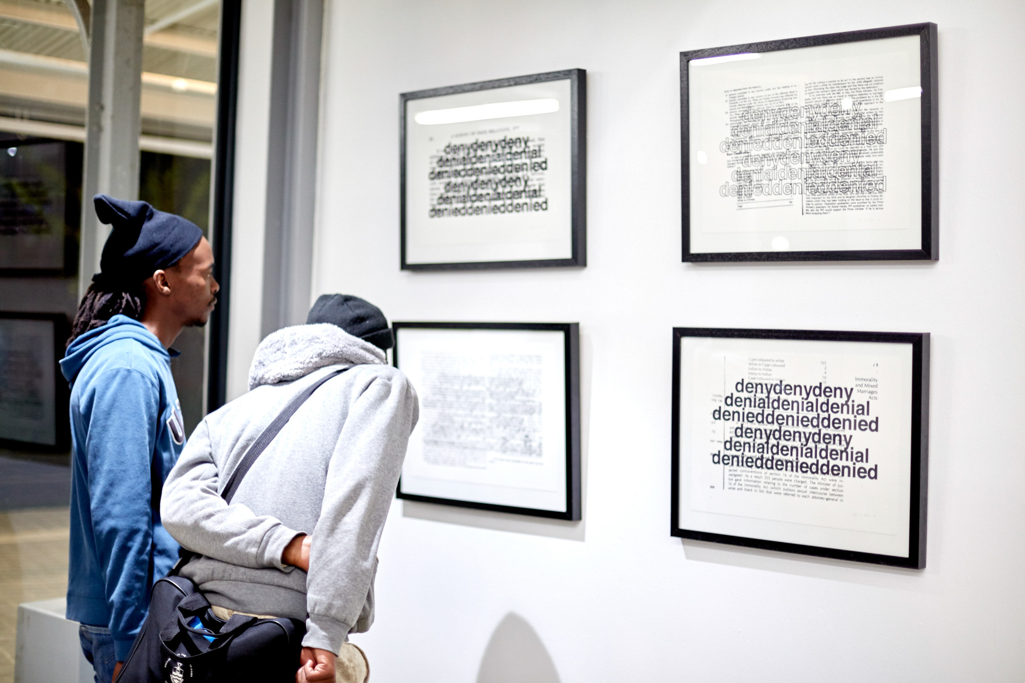 Two African visitors wearing beanies and sweatshirts study four black-framed prints that layer the bold repeated words "deny, denial, denied" over reproduced pages of apartheid-era race legislation.