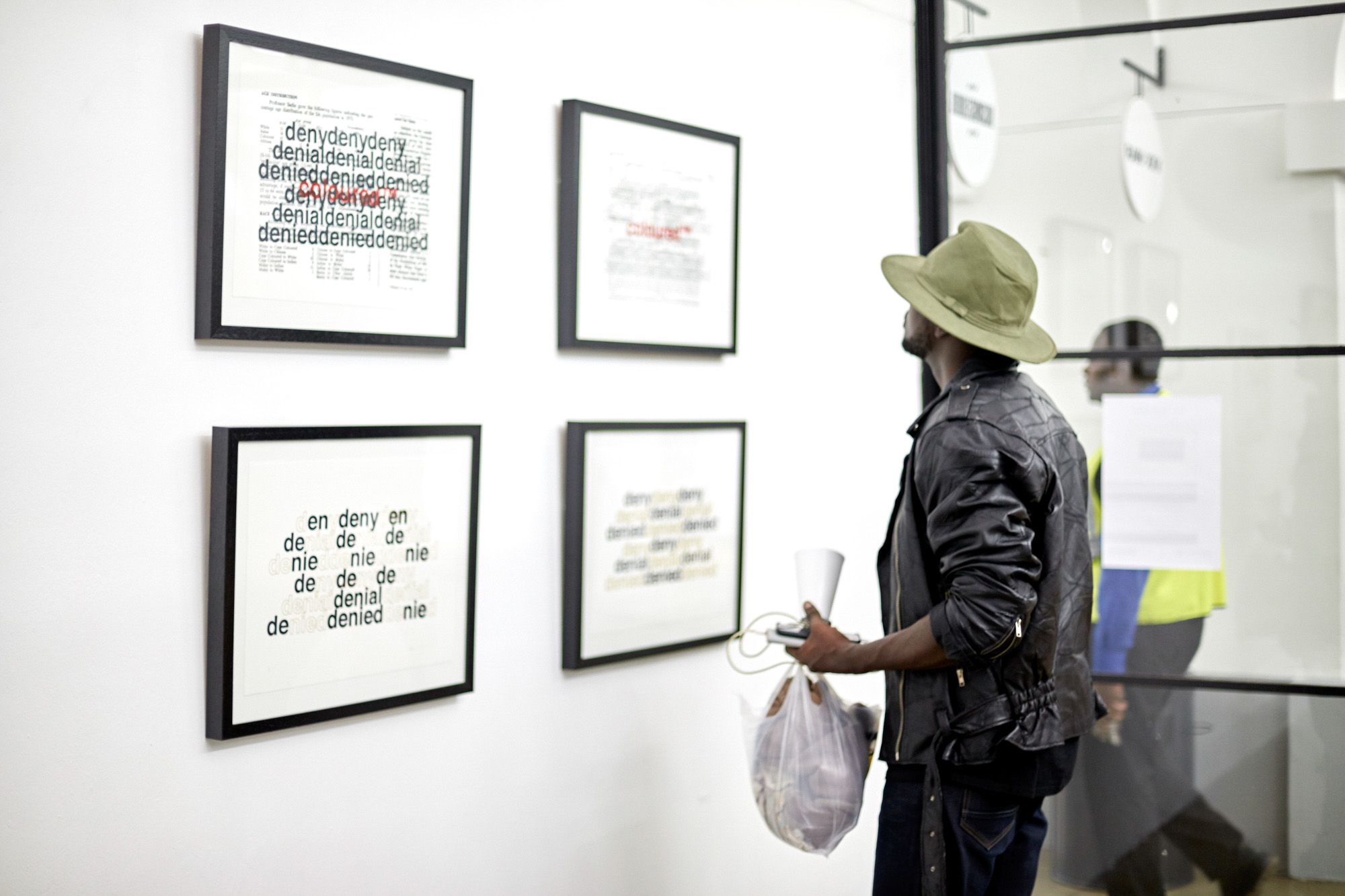 An African male visitor in a leather jacket and wide-brimmed hat looks closely at four framed typographic prints repeating "deny, denial, denied," one overlaid with "coloured™" in red.