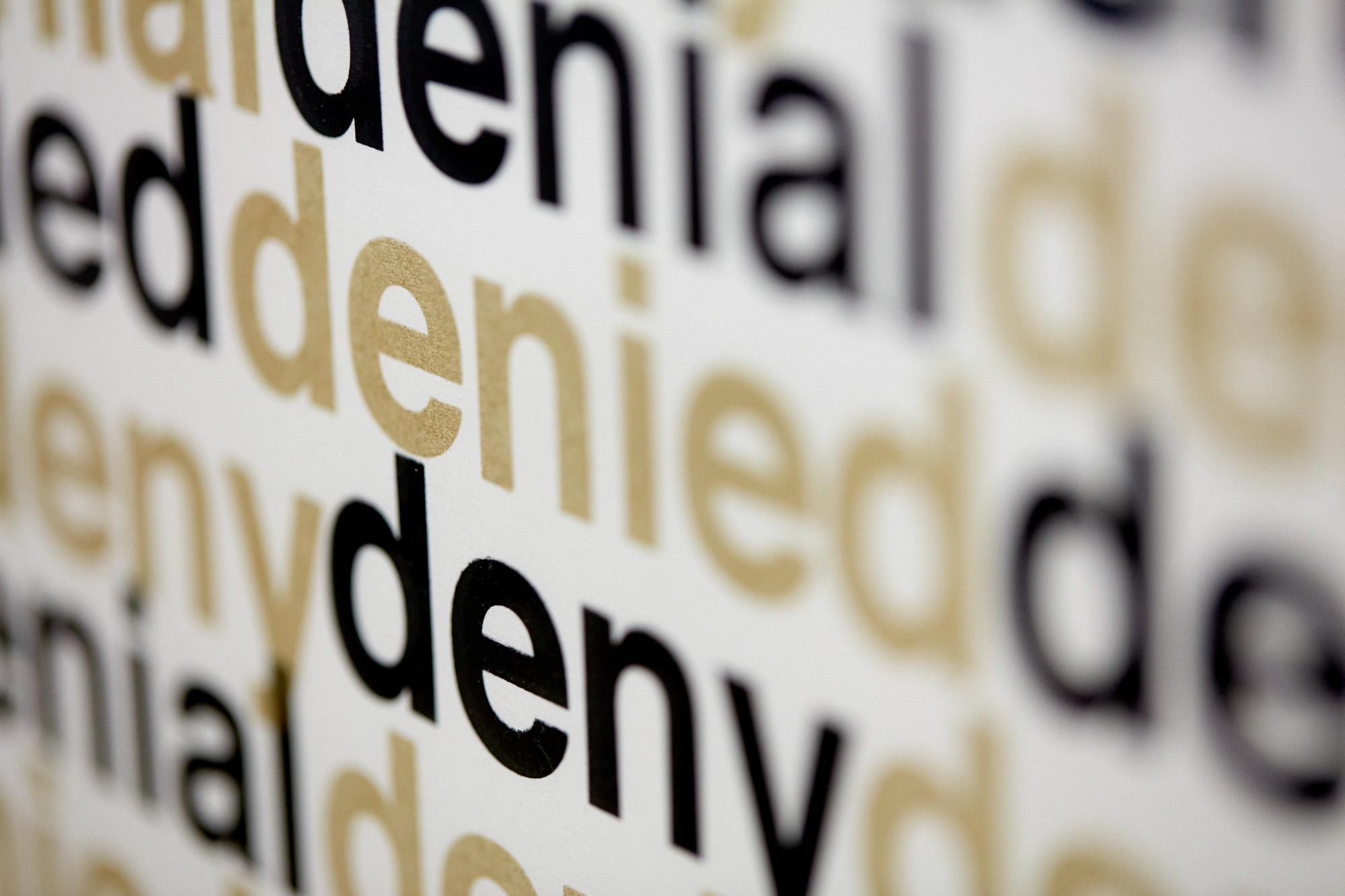 Extreme close-up of a screenprint with the words "deny, denial, denied" repeated in overlapping black and gold lowercase type on white paper, with shallow depth of field.
