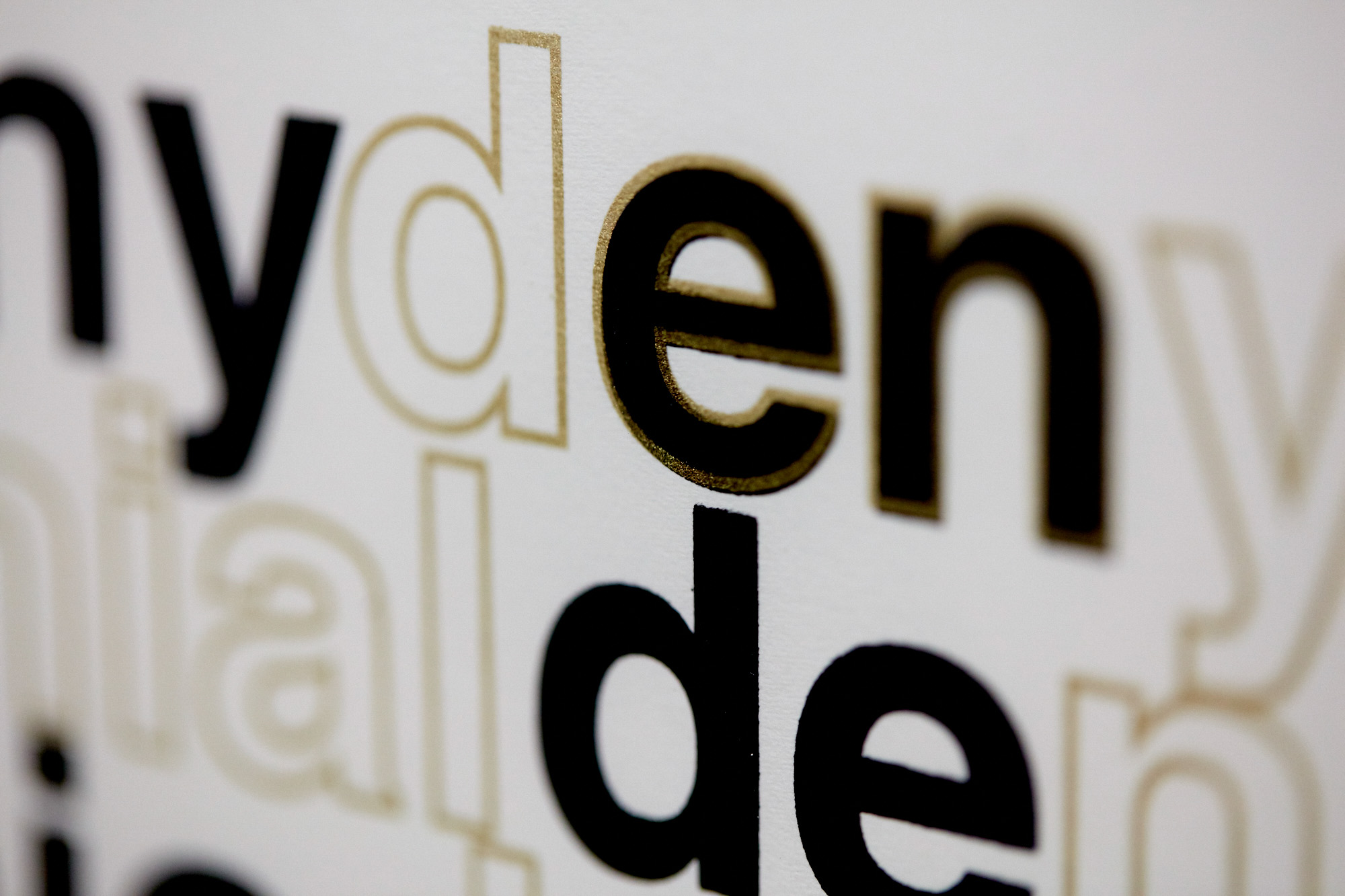 Macro detail of overlapping letters from the words "deny" and "denial" in solid black and outlined metallic gold ink on white paper.